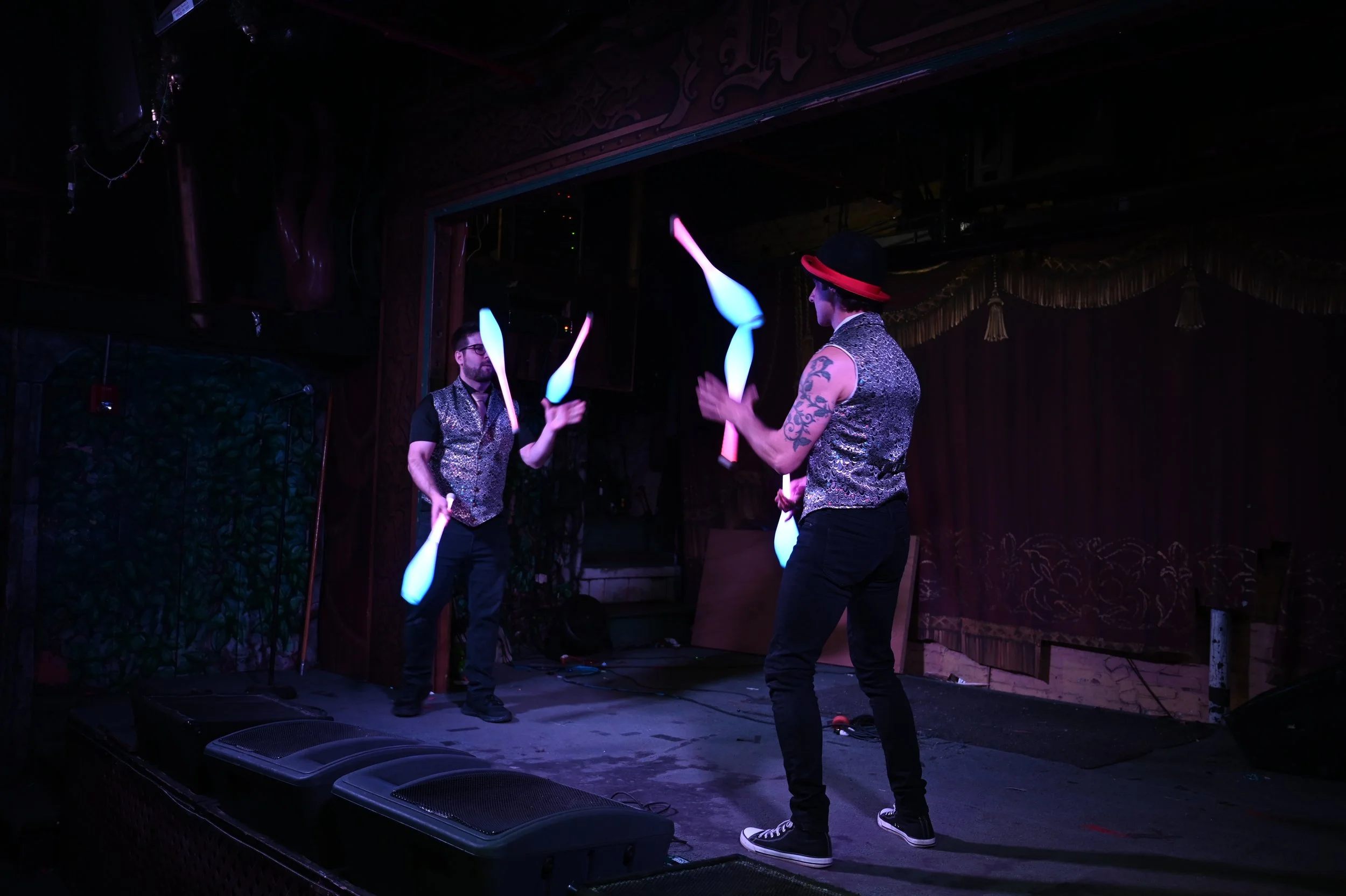 Two performers on stage juggling clubs with glow-in-the-dark blades in a dimly lit venue, wearing similar vests and one wearing a hat.