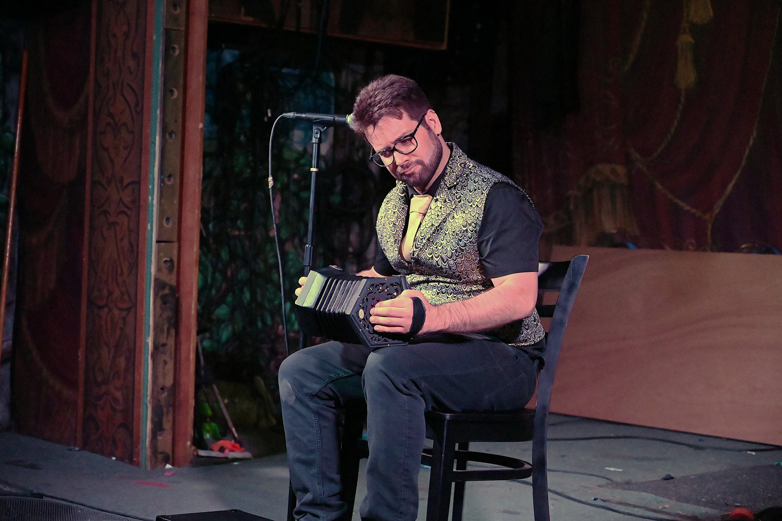 A man wearing glasses and a patterned vest is seated on a chair, playing a concertina on a stage with a microphone nearby.