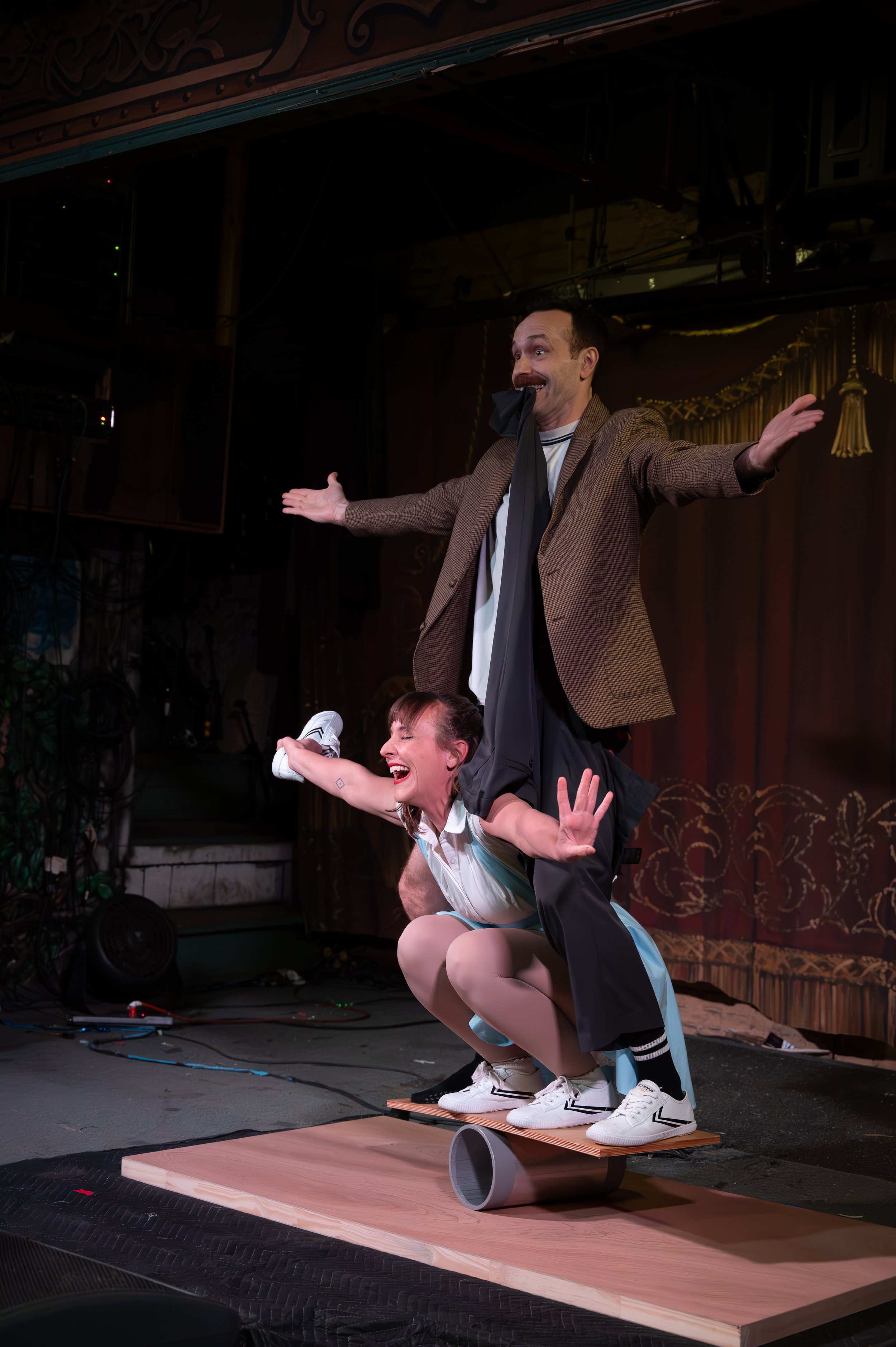 Two performers on stage, a man standing with arms outstretched and a woman crouching beneath him, both smiling and appearing to perform a comedic act. They are on a wooden platform with a pipe underneath and a theatrical curtain in the background.