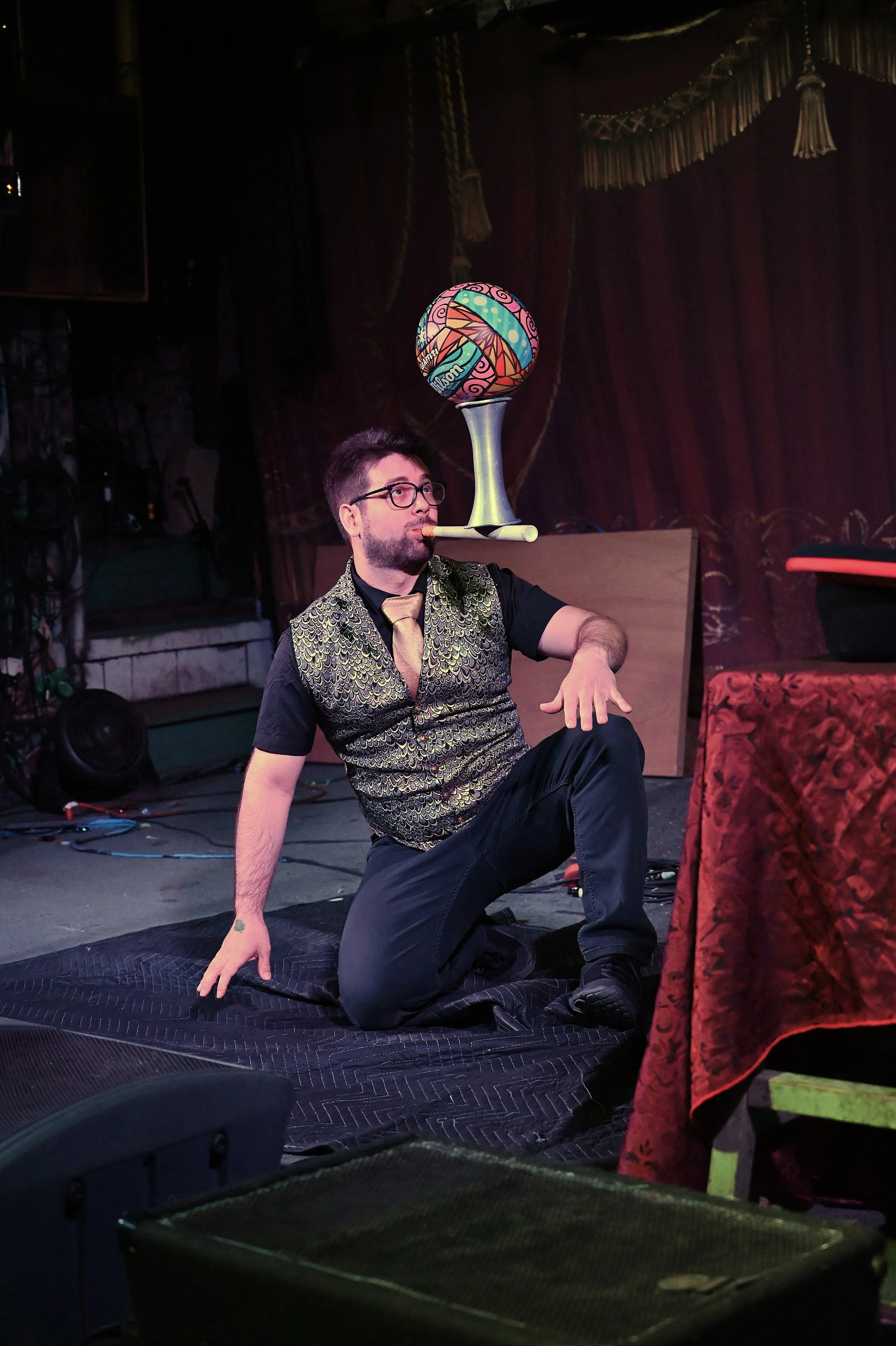 A performer balancing a decorative ball on a pedestal balanced on his nose while sitting on a stage floor.