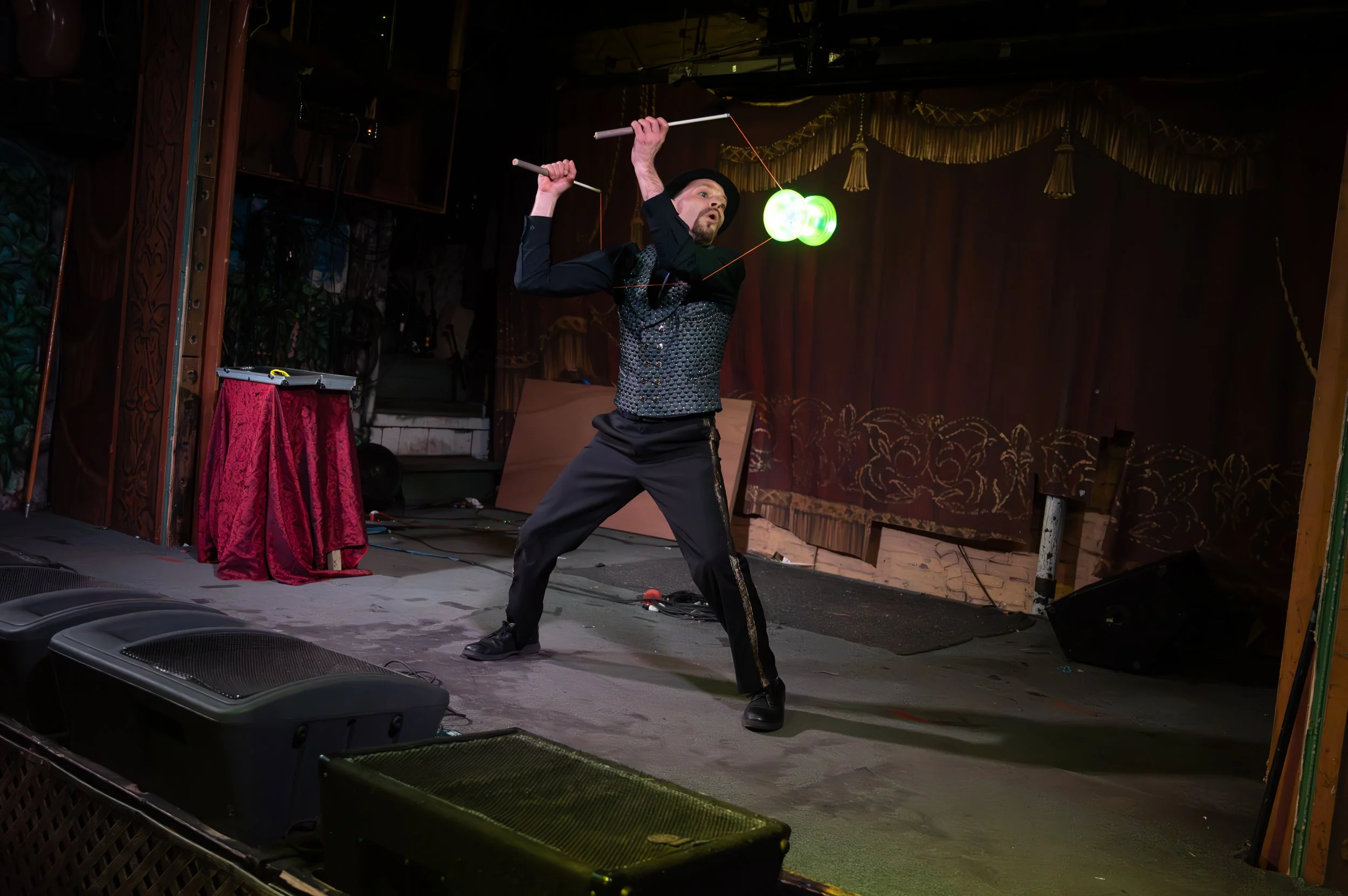 Performer on stage, wearing a black hat and vest, manipulating glow balls with poles in a theatrical setting.