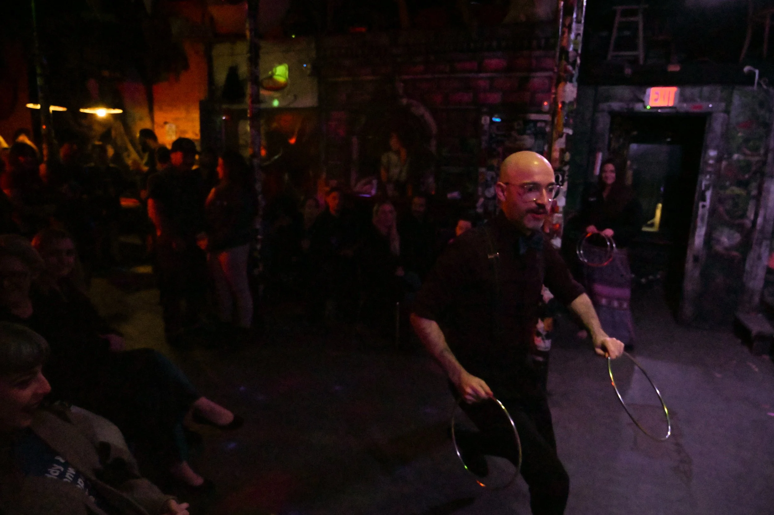 A bald man with glasses holding two hula hoops in a dimly lit, crowded indoor venue with exposed brick walls and vintage decor. There are people sitting and standing, watching him perform.