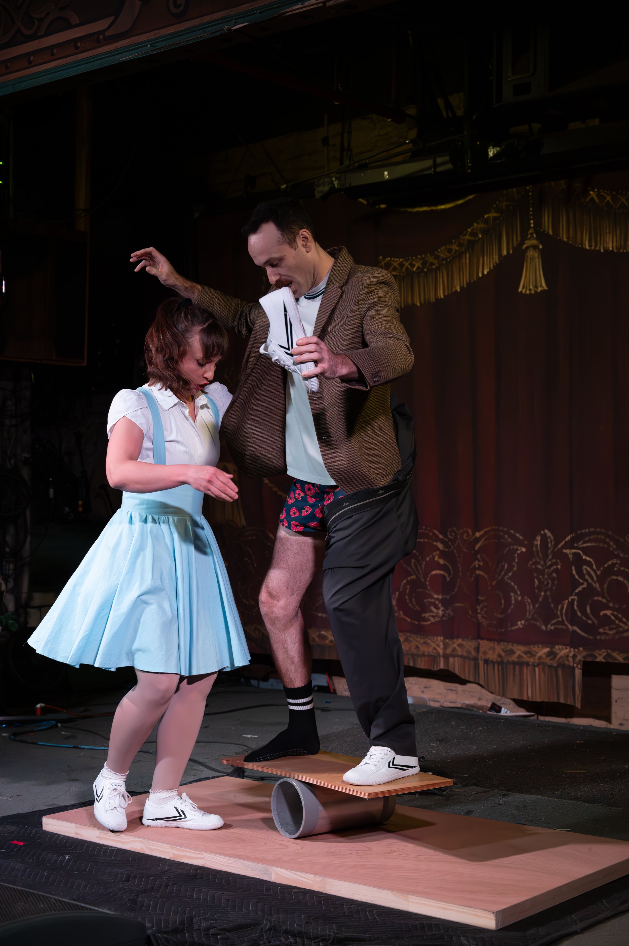 A man and a woman are on a stage, with the man balancing on a wooden plank supported by a cylindrical object, holding a shoe, while the woman looks closely at his leg. The scene appears comedic or theatrical.