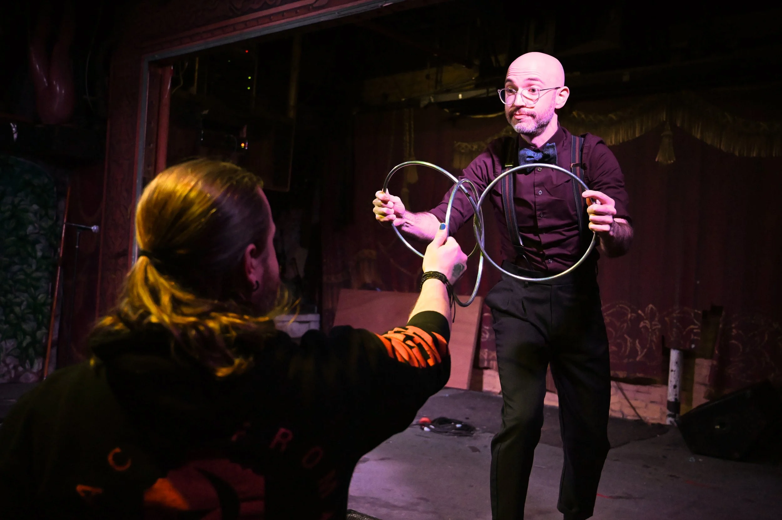 A magician performing a trick with metal hoops on stage, with a woman in the audience holding one of the hoops. The stage has dark curtains and red lighting.