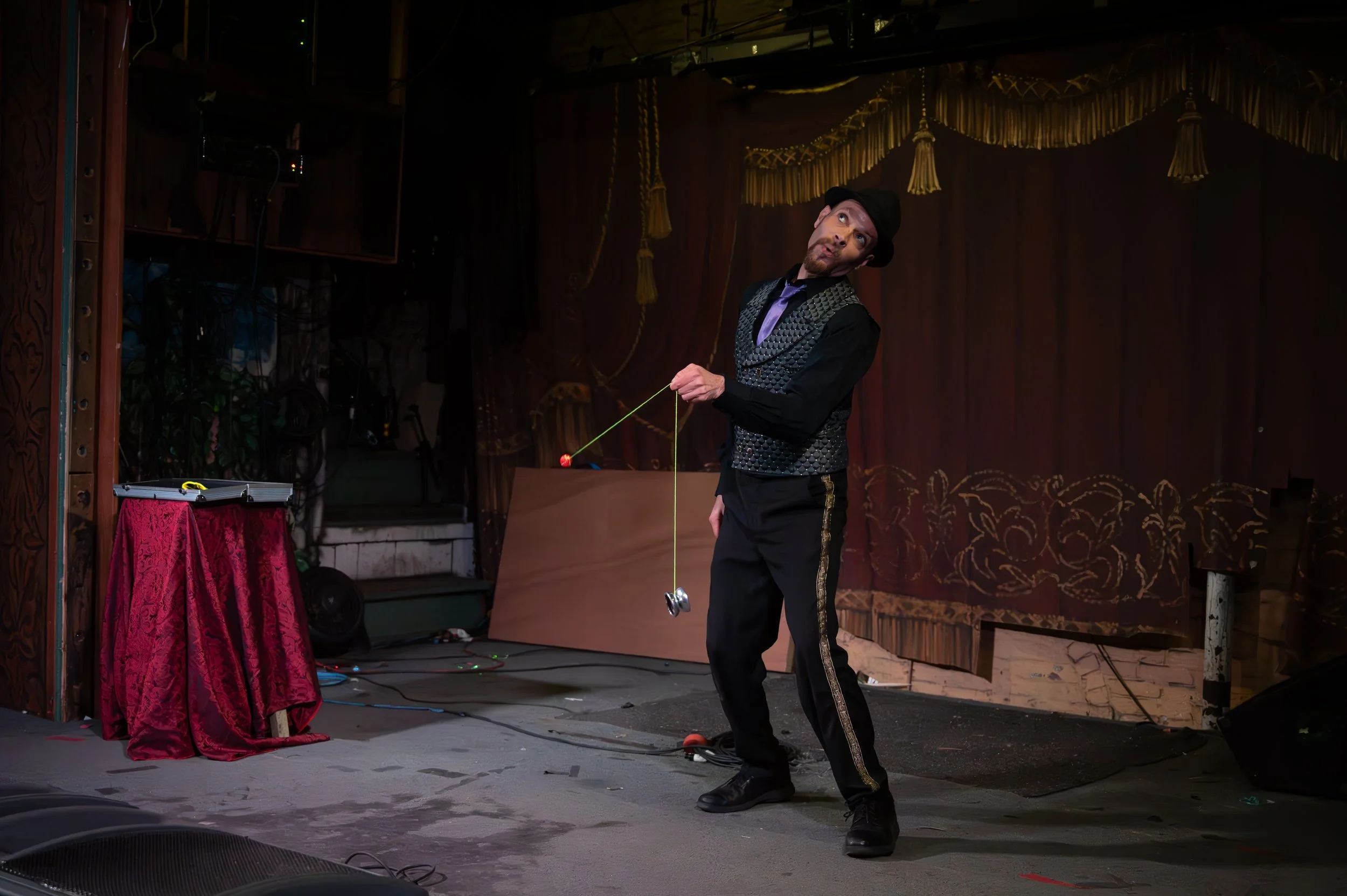 Performer on stage in a black costume with patterned vest and bowler hat, spinning a clown yo-yo, stage with red curtains and a small table with a red cloth.