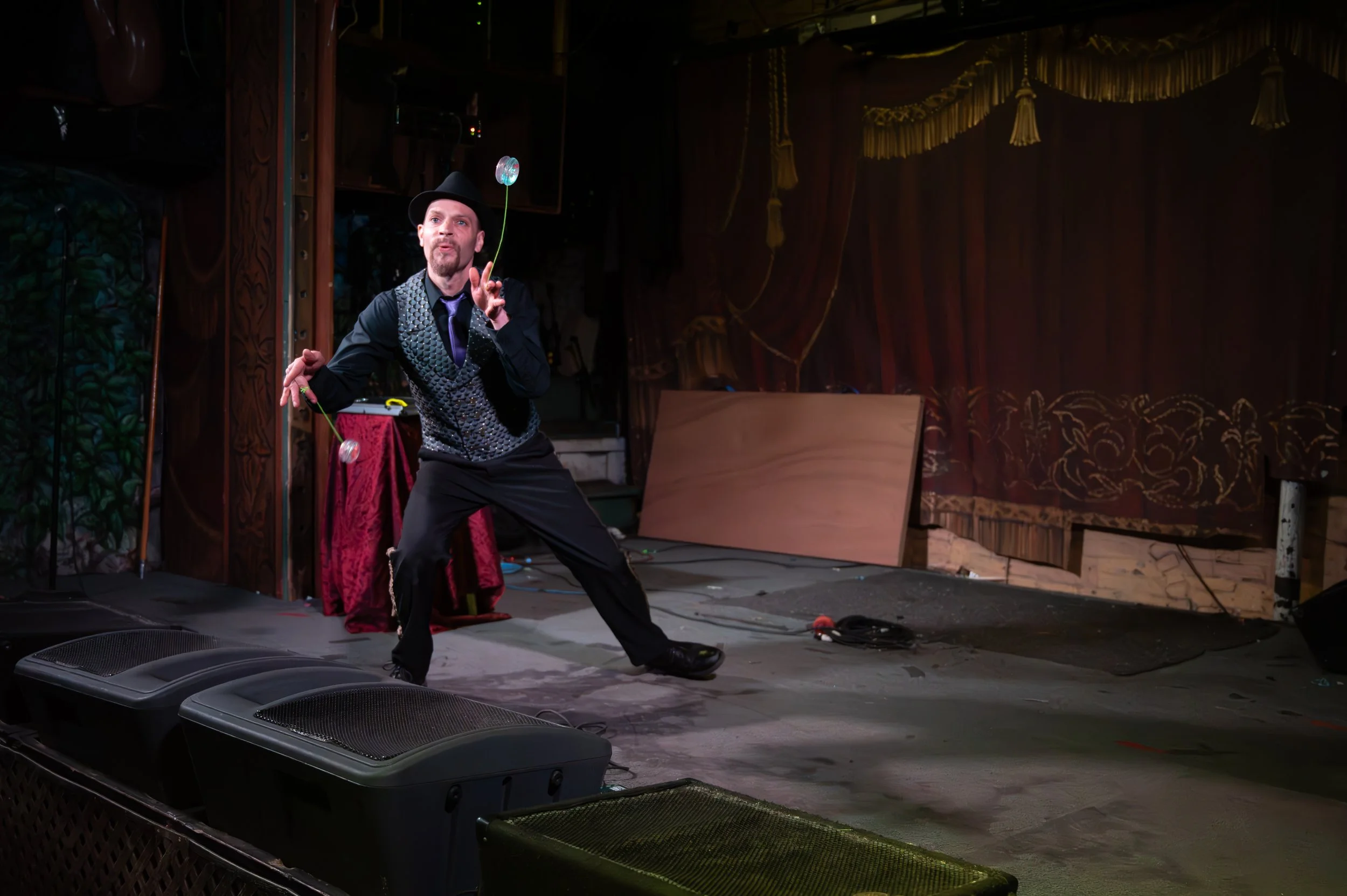 A man performing a juggling act on stage with a dark background, wearing a black hat, black shirt, and a shiny vest, with red curtains behind him and stage speakers in the foreground.