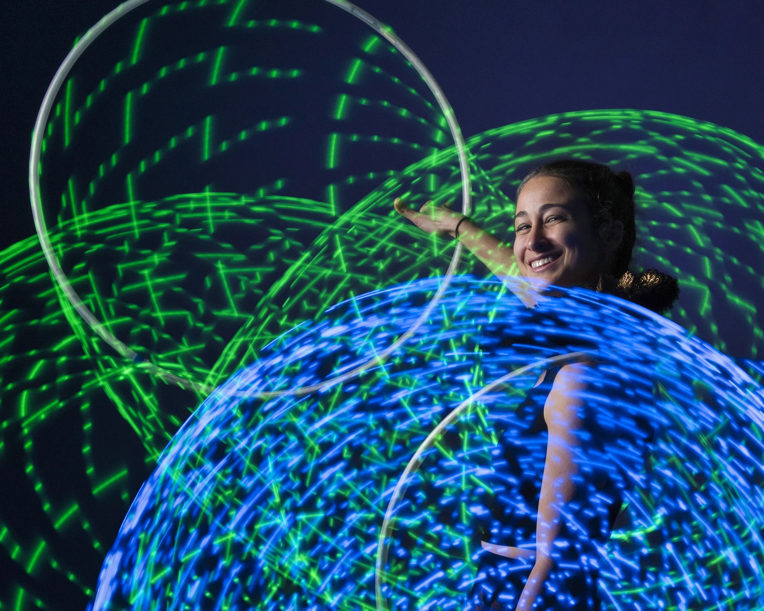 A smiling woman interacts with colorful light trails created by long exposure photography, featuring green and blue glowing orbs and streaks in front of a dark background.