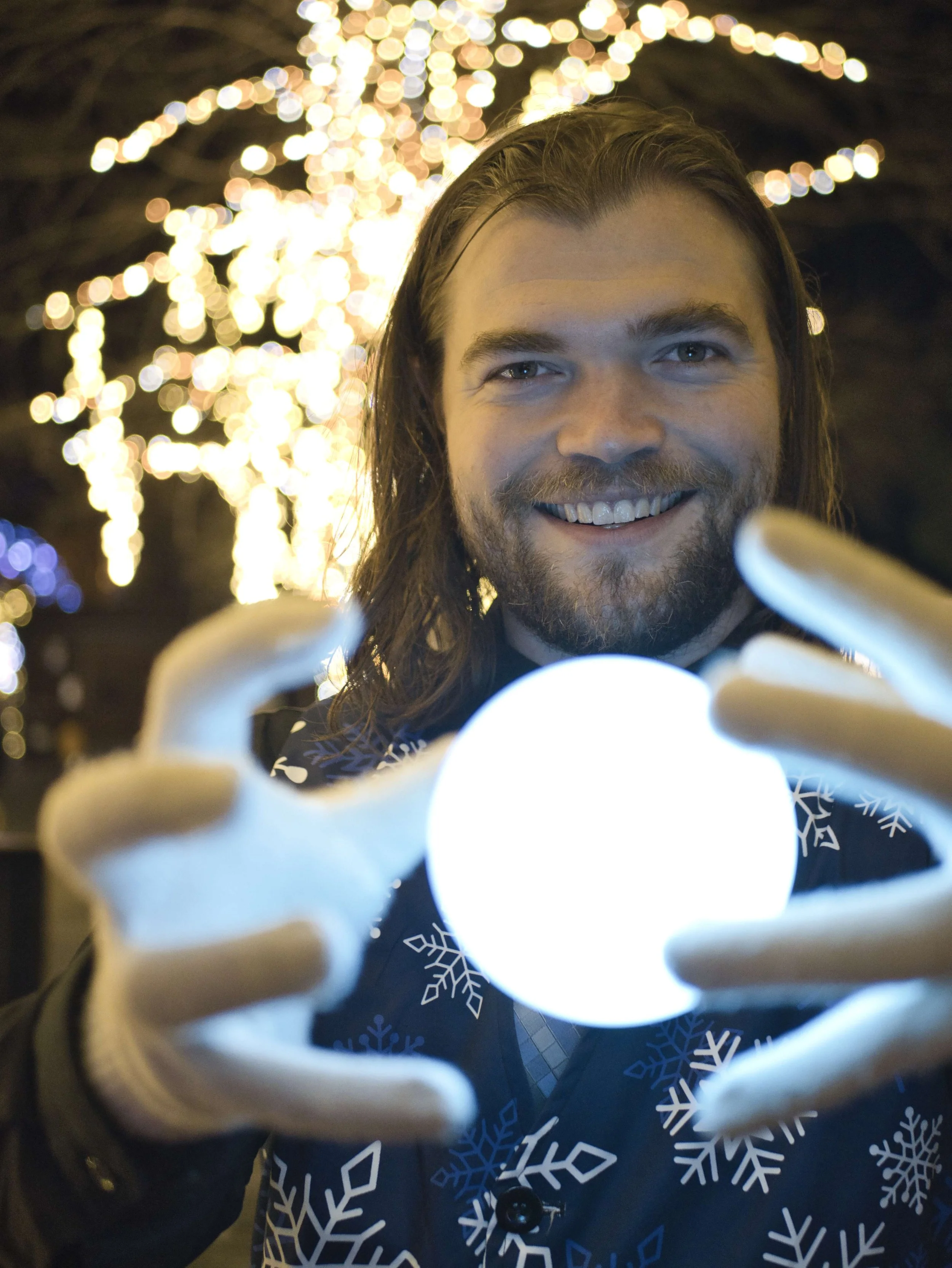 A man with long hair and a beard holding a glowing orb at night, with Christmas lights in the background.