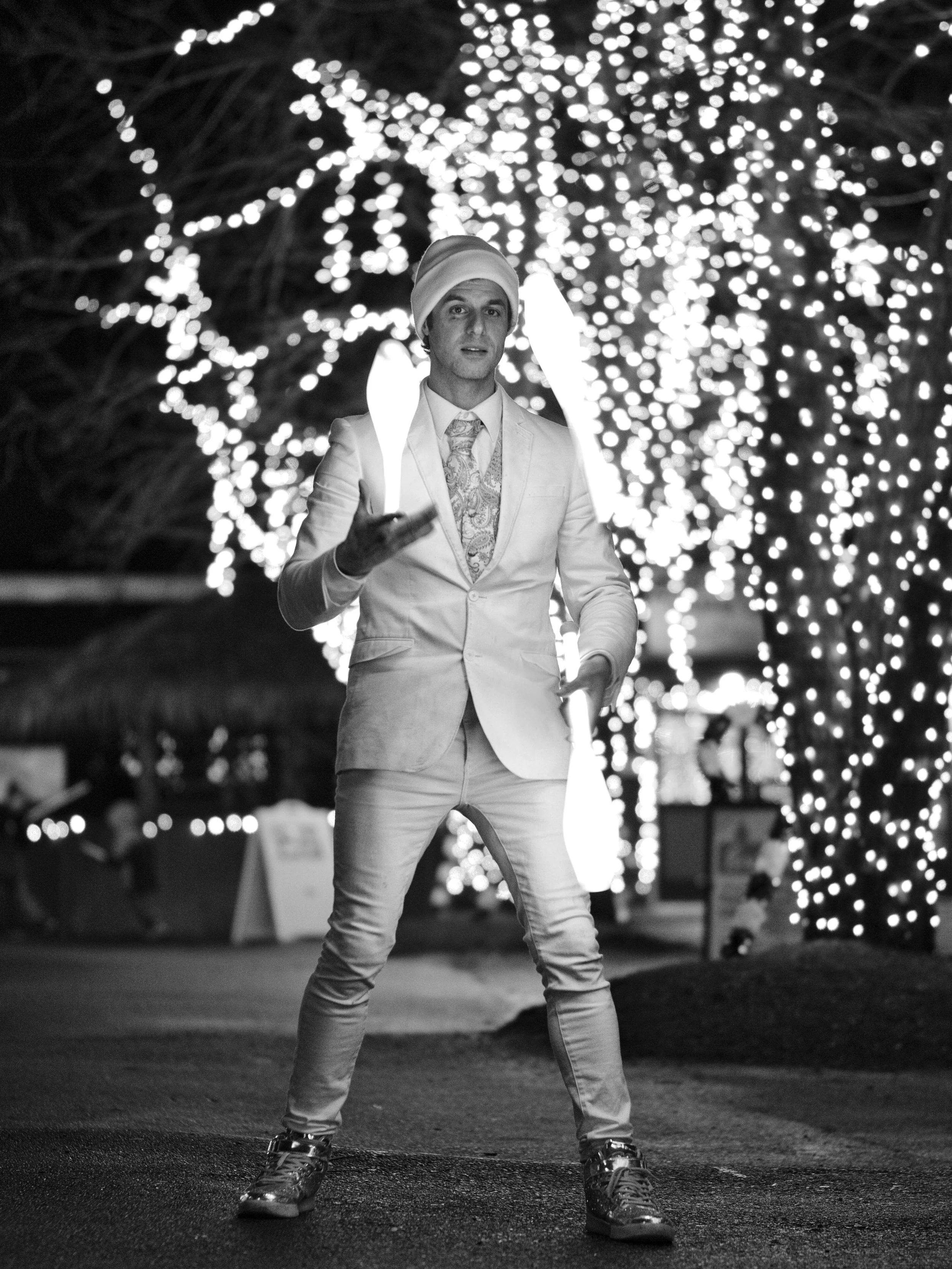 A man dressed in a suit and tie, wearing a beanie, stands outdoors at night in front of a tree decorated with bright string lights. He is making a gesture with his right hand.