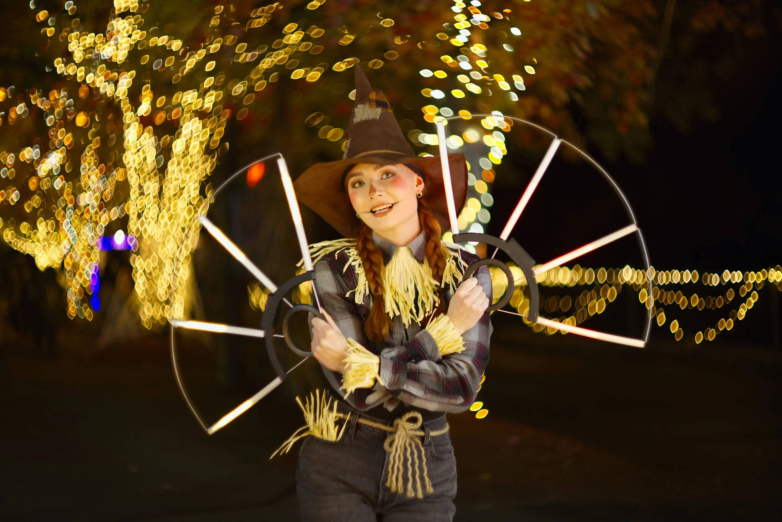 A woman dressed as a scarecrow holding a light-up wheel prop during a nighttime outdoor event, with blurred yellow Christmas lights in the background.