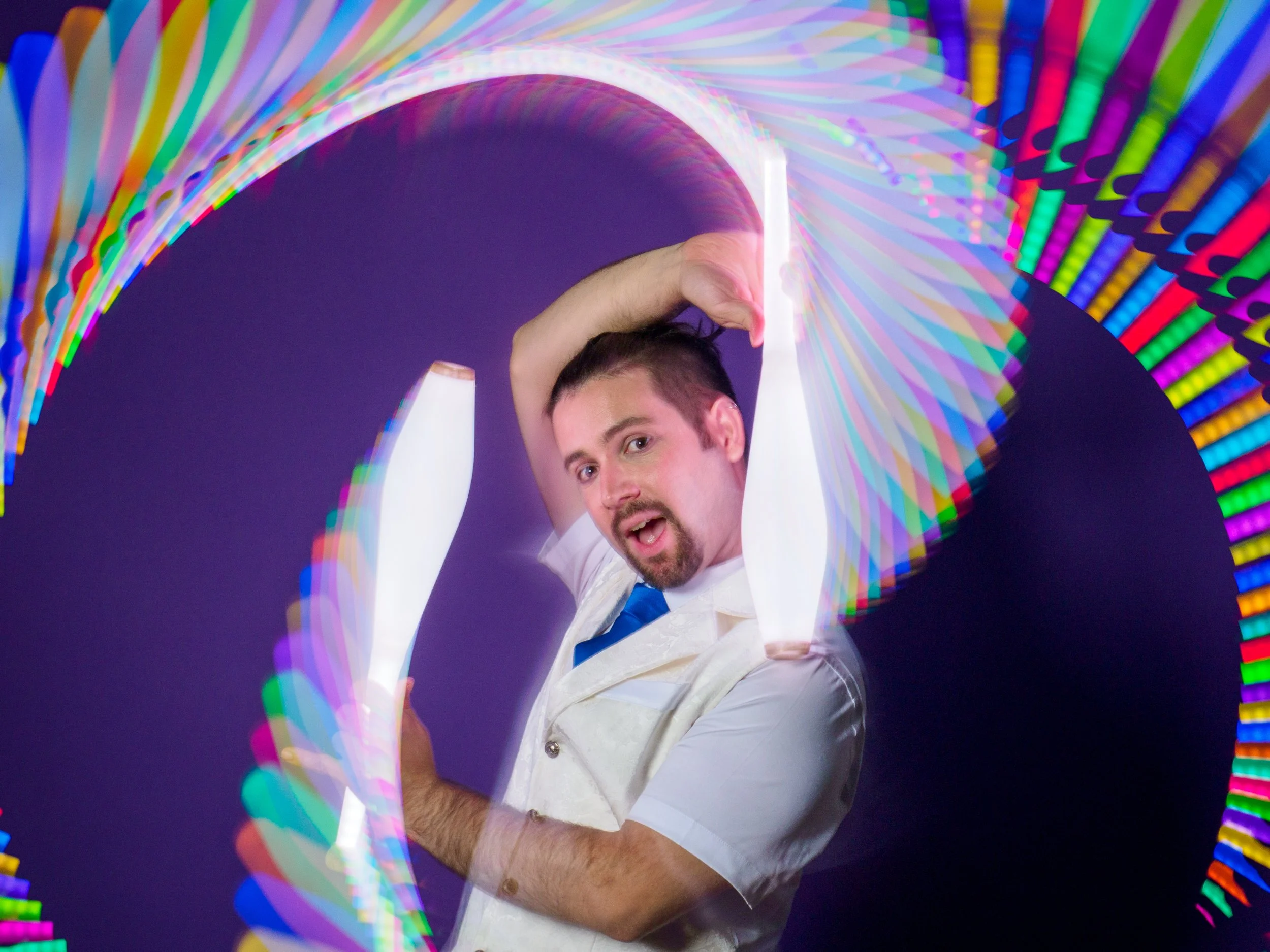 A man with short dark hair and a beard, wearing a white shirt and blue tie, stands in front of a dark background, holding colorful light tubes that create a rainbow-like swirling pattern around him.