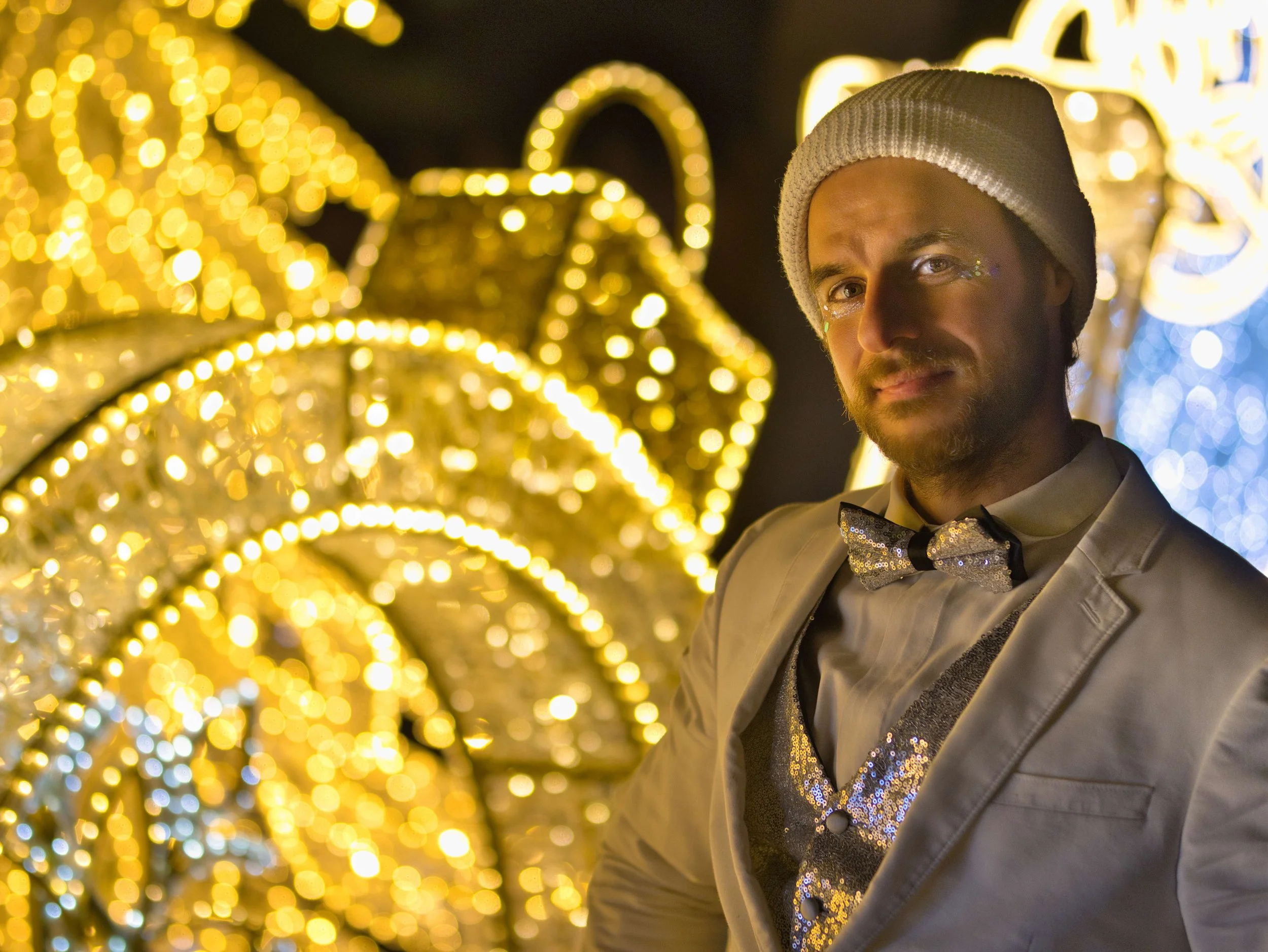 A man dressed in a silver suit with a shiny bow tie, wearing a beanie, standing in front of illuminated decorative arches at night, with festive holiday lights creating a bokeh background.