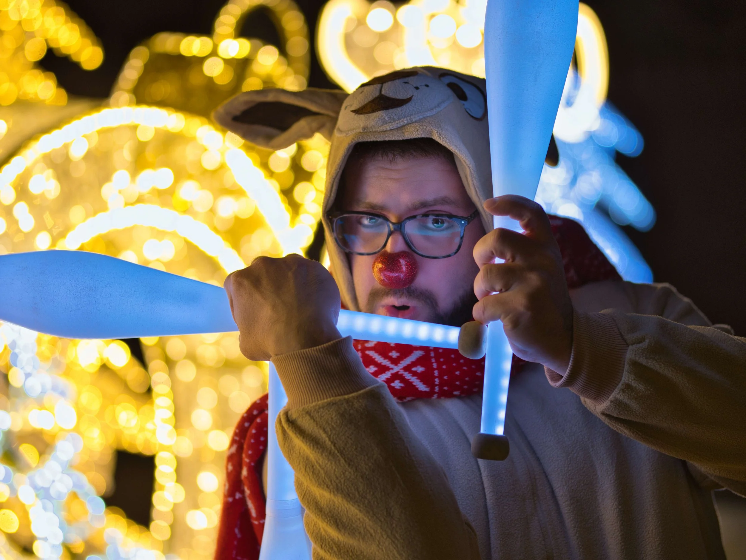 Man dressed as a reindeer with a red nose, glasses, and a hat with reindeer face, holding illuminated light sticks, surrounded by Christmas lights.