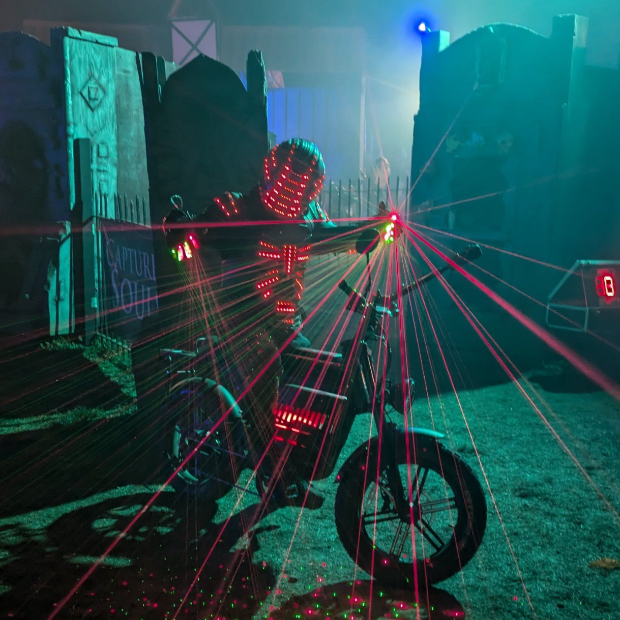 A person dressed as a robot with LED lights on a bicycle, surrounded by colorful laser beams in a dark setting.
