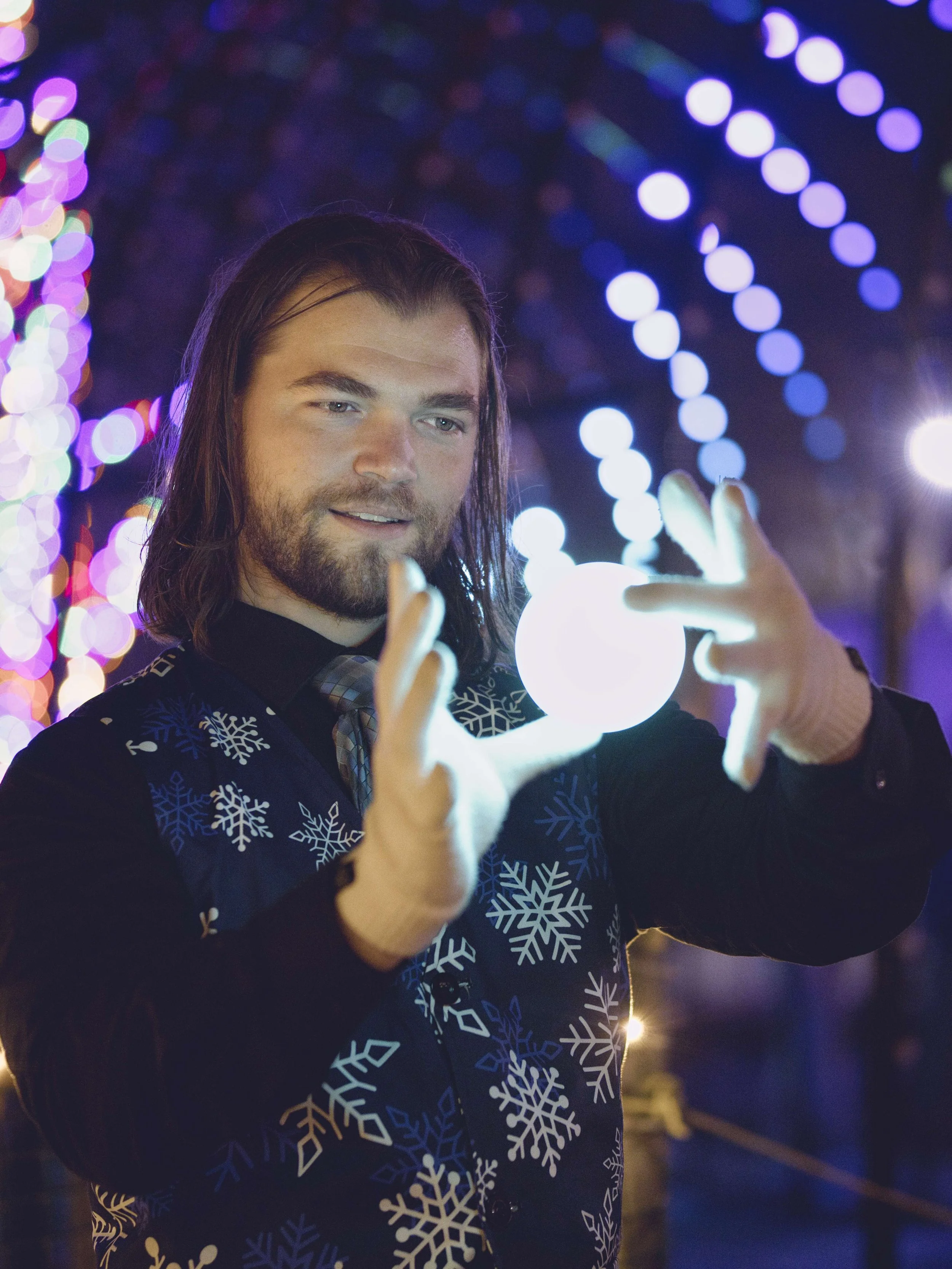 A man with long hair and a beard is holding a glowing spherical object at night during a festive event. He is dressed in a dark sweater with white snowflake patterns, and the background is decorated with colorful, blurred holiday lights.