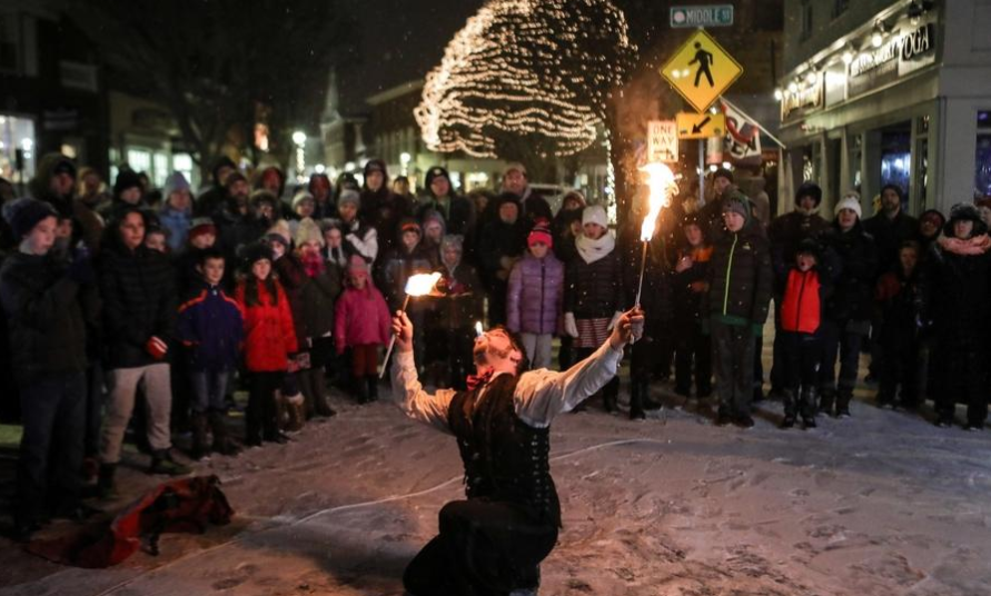 A fire eater, holding fire torches, kneels in the snow during a nighttime event. A crowd of people, dressed warmly, surrounds the performer, watching attentively. Christmas lights are visible in the background.