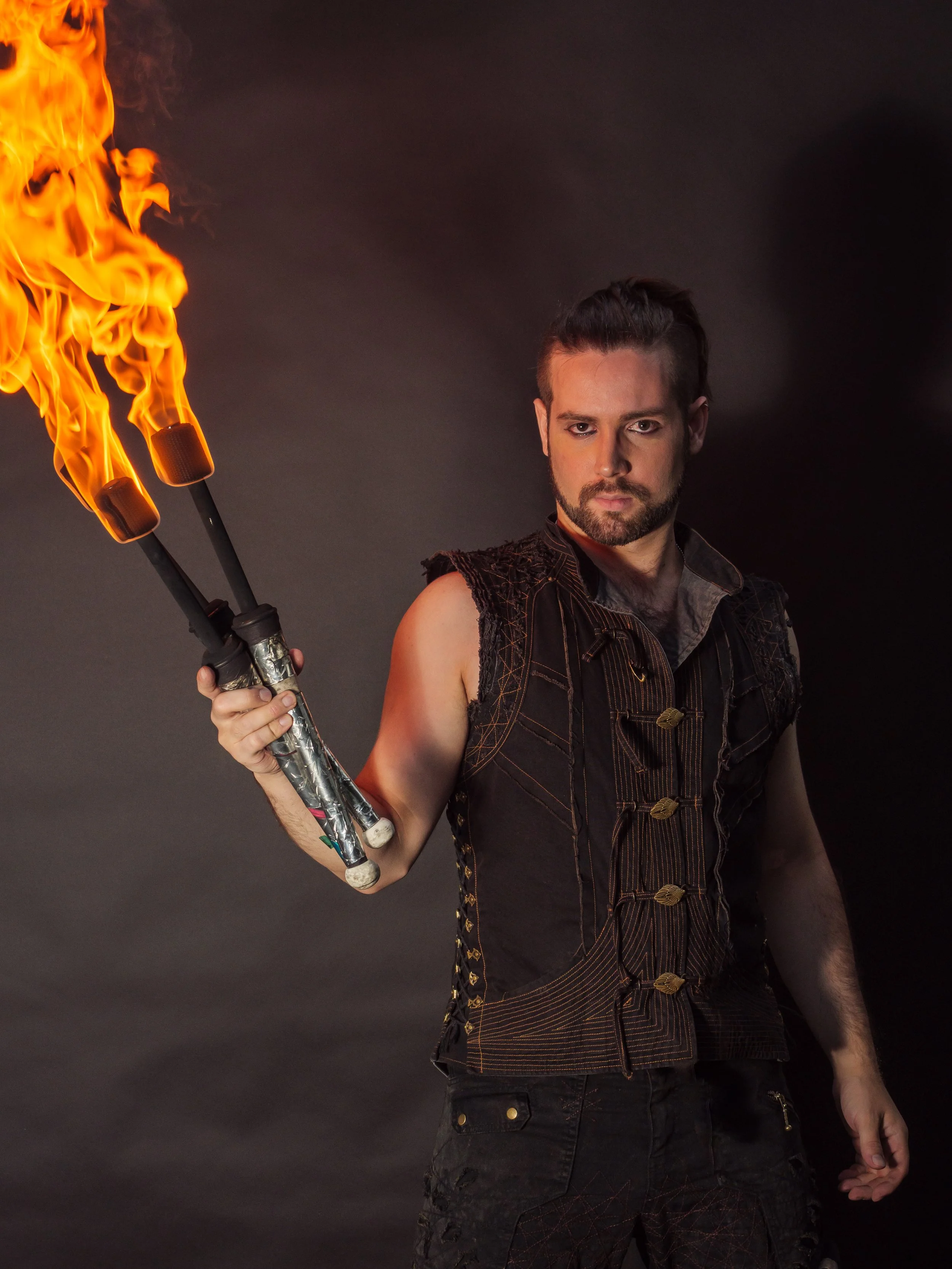A man with dark hair and a beard holding a flame juggling torch against a black background.