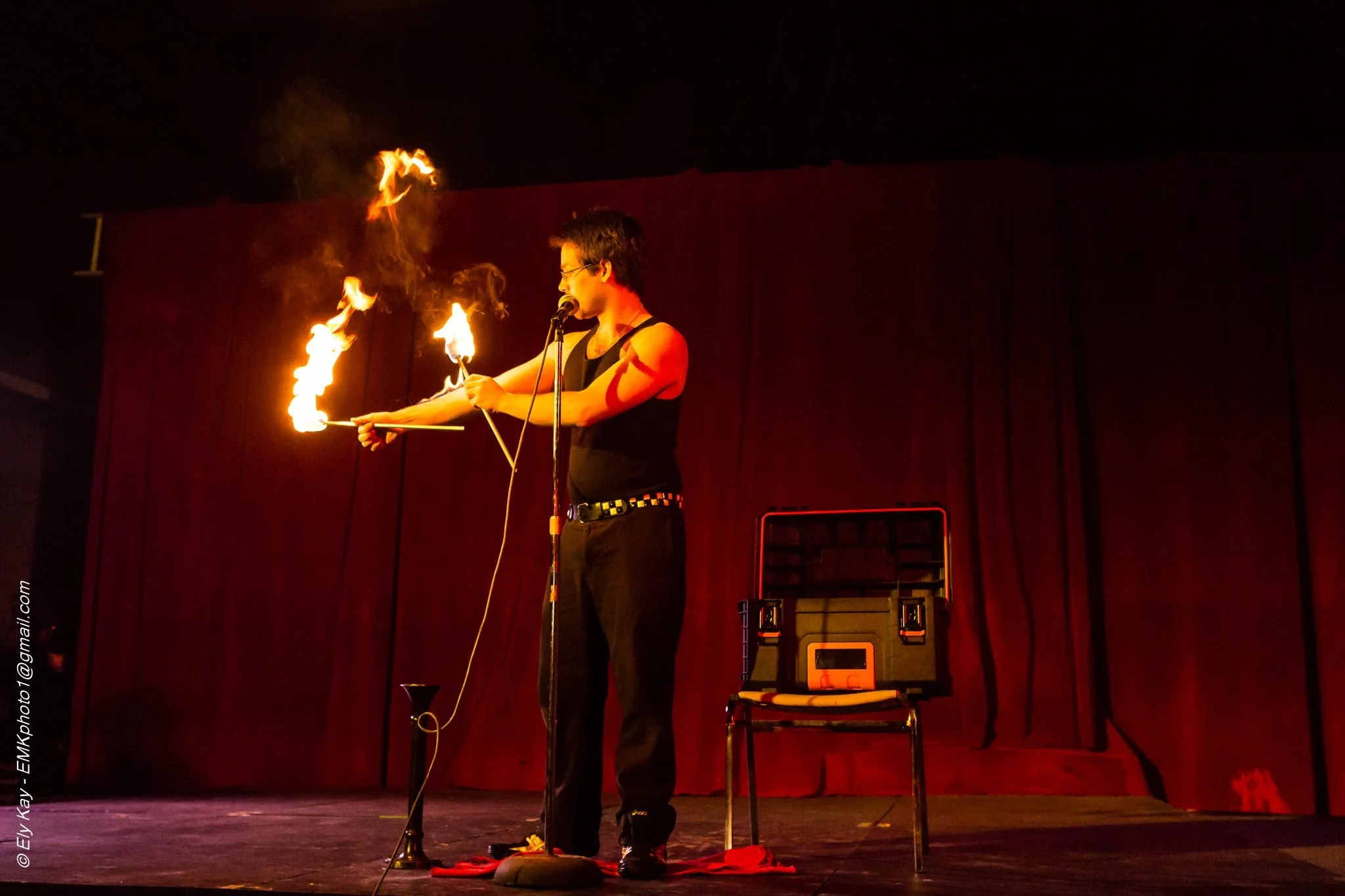 A performer on stage with a red curtain background demonstrating fire juggling, holding lit torches, and wearing a black sleeveless top and black pants.