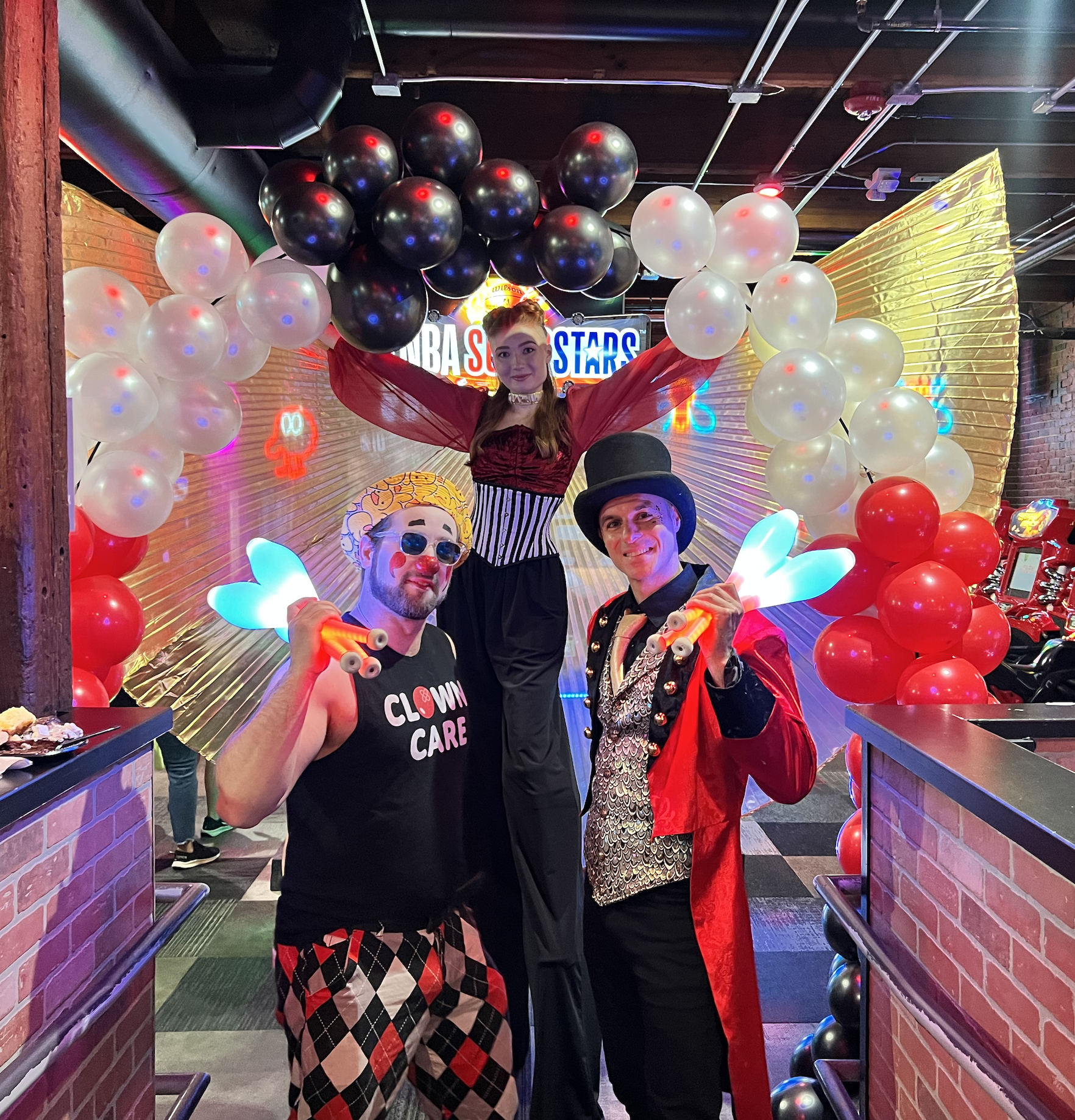 Two performers in circus costumes pose in front of a woman on stilts at a circus-themed event, with colorful balloons and a neon sign in the background.