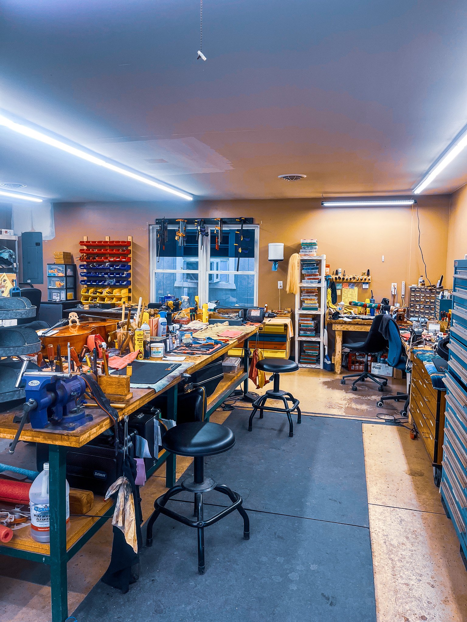 View of a cluttered workshop with workbenches, tools, and shelving units filled with supplies and equipment.