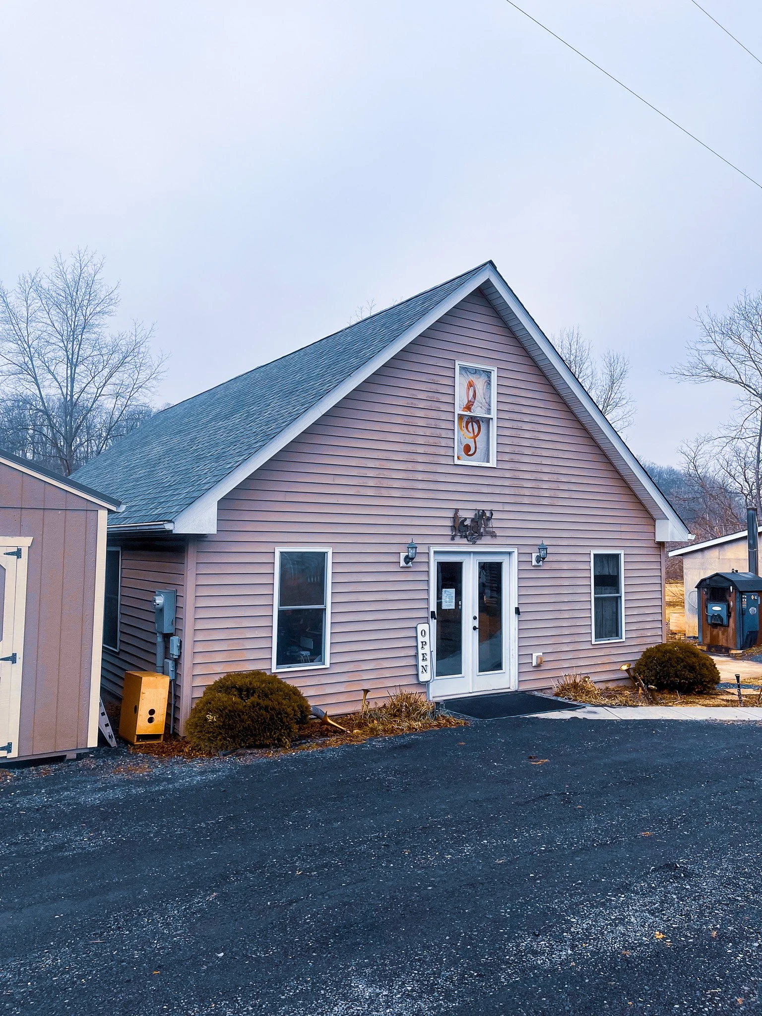 A pink building with a white double door, surrounded by bushes, with a sign that reads 'OPEN' near the door. The building has a musical note decoration above the door and a treble clef window at the top. There is outdoor lighting and a mailbox to the right.