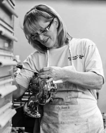 A woman with glasses working on a mechanical device in a workshop, wearing an apron that says "My Aunt B."