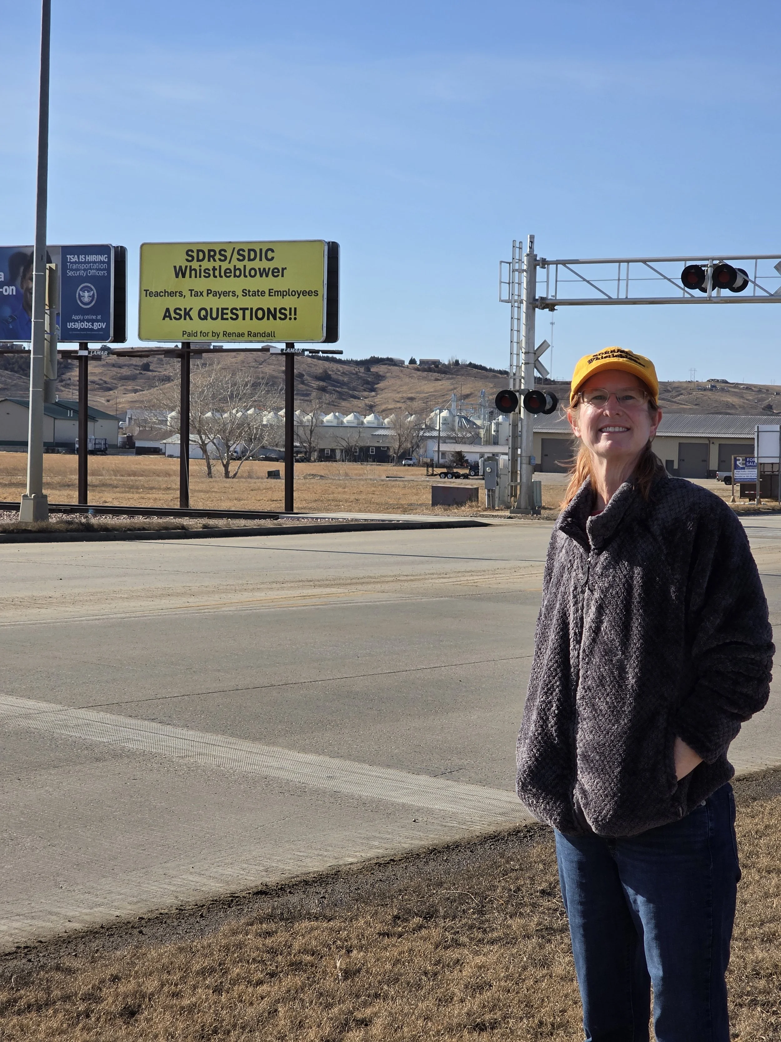 Ft. Pierre large yellow sign in the background that reads, 'SDRS/SDIC Whistleblower Teachers, Tax Payers, State Employees ASK QUESTIONS!! Paid for by Renae Randall'.