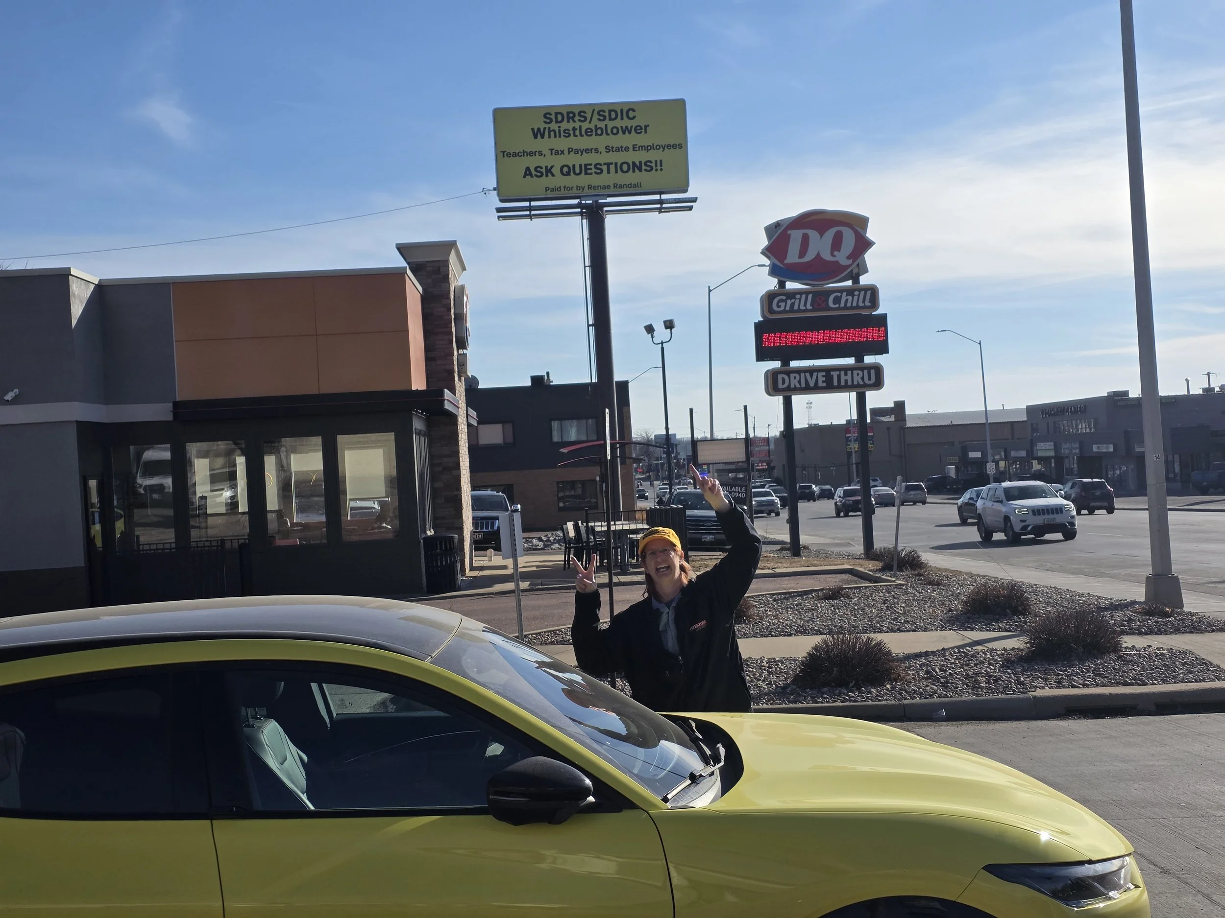 Sioux Falls large yellow sign in the background that reads, 'SDRS/SDIC Whistleblower Teachers, Tax Payers, State Employees ASK QUESTIONS!! Paid for by Renae Randall'.