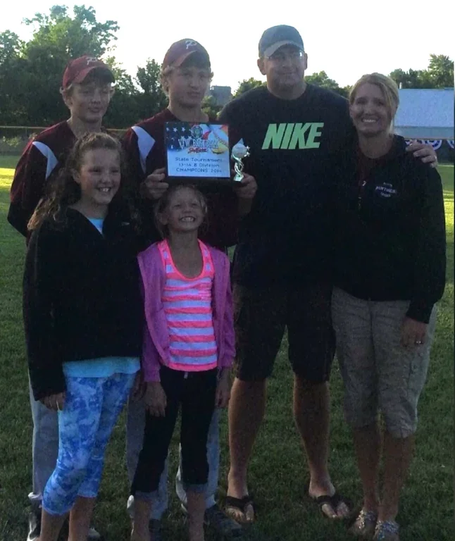 Group of six people, including children and adults, standing on a grassy field during daytime, celebrating a sports event or tournament. One girl is holding a certificate and a small trophy. Everyone is smiling and dressed casually, with trees and a blue sky in the background.