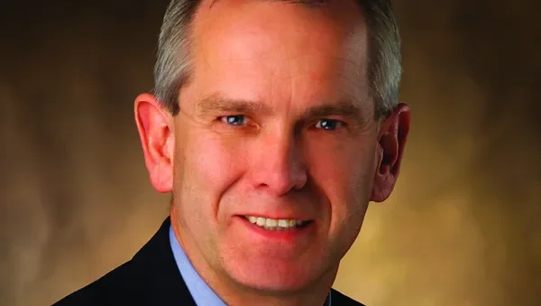 Close-up portrait of a smiling middle-aged man with short gray hair, wearing a dark suit and light blue shirt.