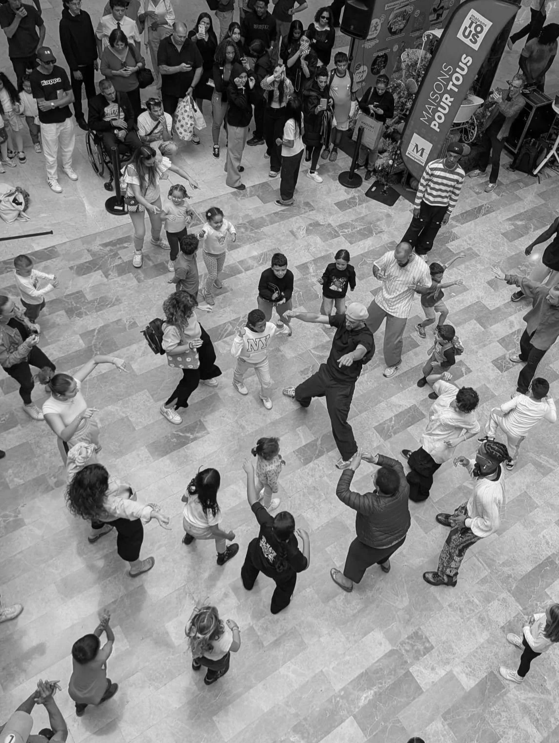 Un groupe de personnes, adultes et enfants, dansant au centre d'un espace public, probablement dans un centre commercial, avec une foule regardant autour d'eux.