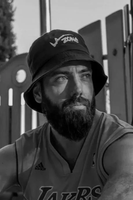 Un homme avec une barbe portant un chapeau noir avec le logo 'VZONE' et un maillot des Los Angeles Lakers, assis à côté d'une clôture en bois, regardant à gauche.