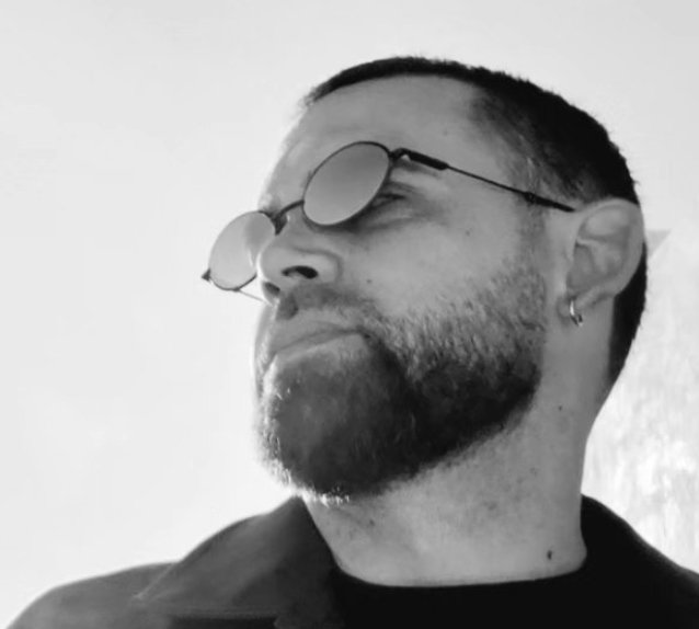 Homme portant des lunettes rondes et une barbe, regardant vers la droite en plein air.