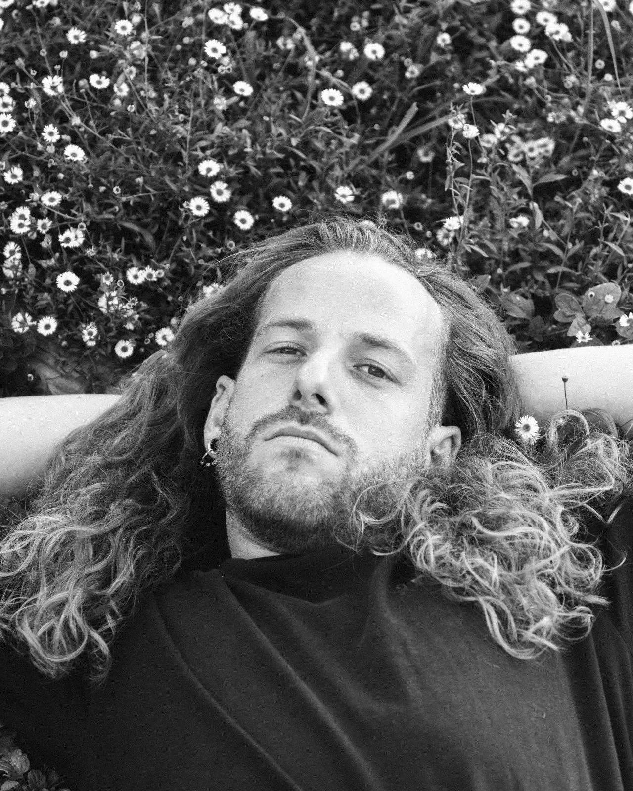 Un homme à cheveux longs et barbe, penchant la tête arrière, allongé dans un champ de petites fleurs, en noir et blanc.
