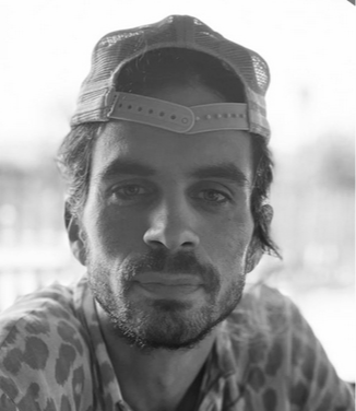 Un homme avec une casquette en arrière, barbe et vêtements à motifs, regardant vers l'appareil photo.