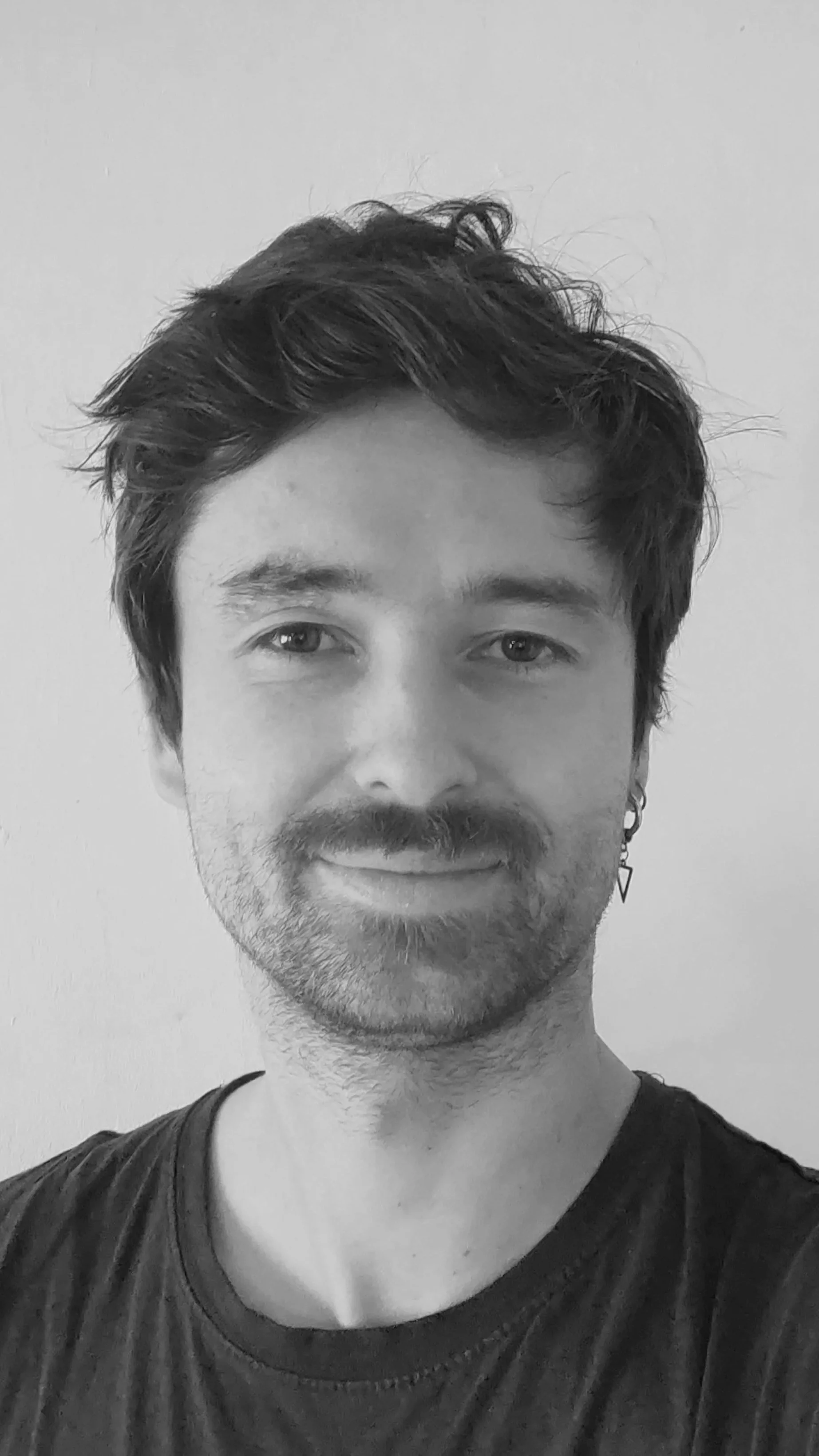 Un portrait noir et blanc d'un homme avec des cheveux bouclés, une barbe et une boucle d'oreille en forme de triangle, portant un t-shirt.