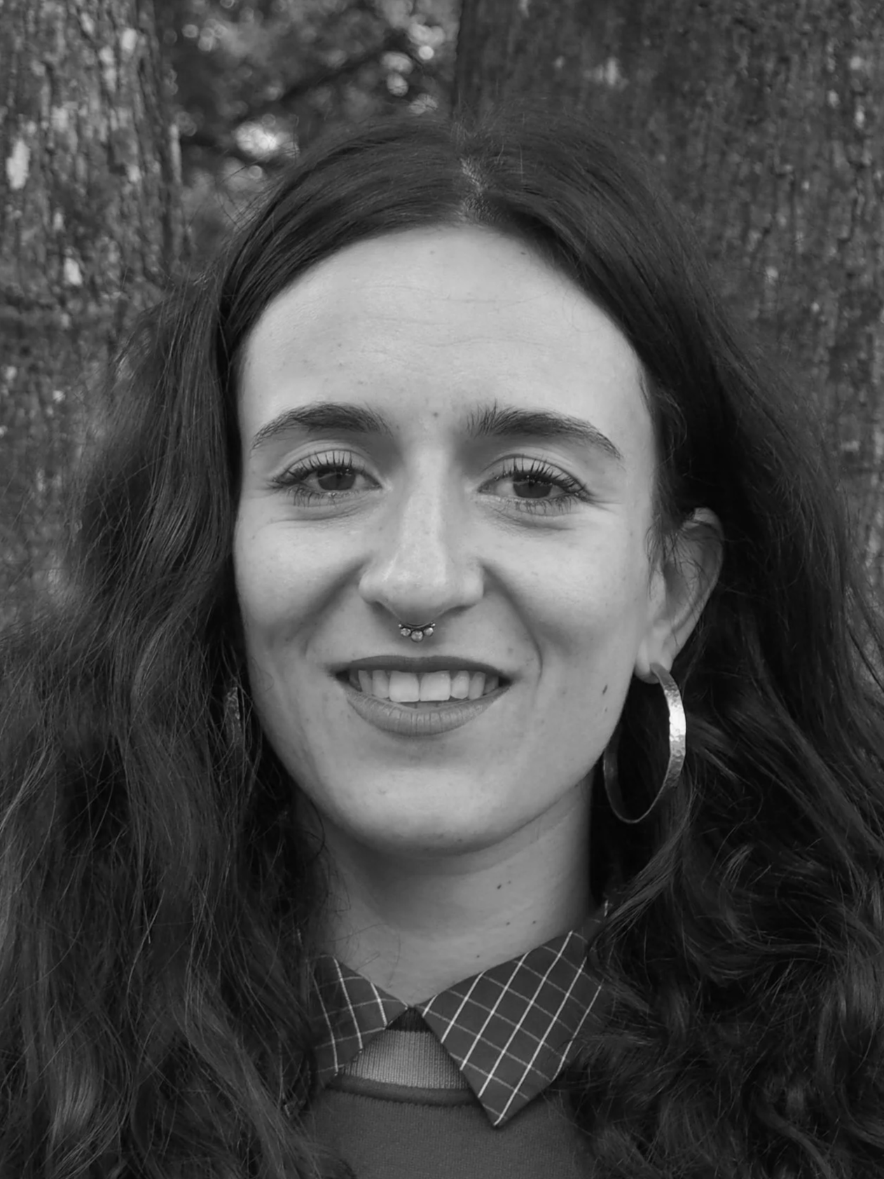Portrait en noir et blanc d'une jeune femme souriante avec des cheveux longs et ondulés, portant une boucle d'oreille et un piercing septum, en extérieur avec des arbres en arrière-plan.