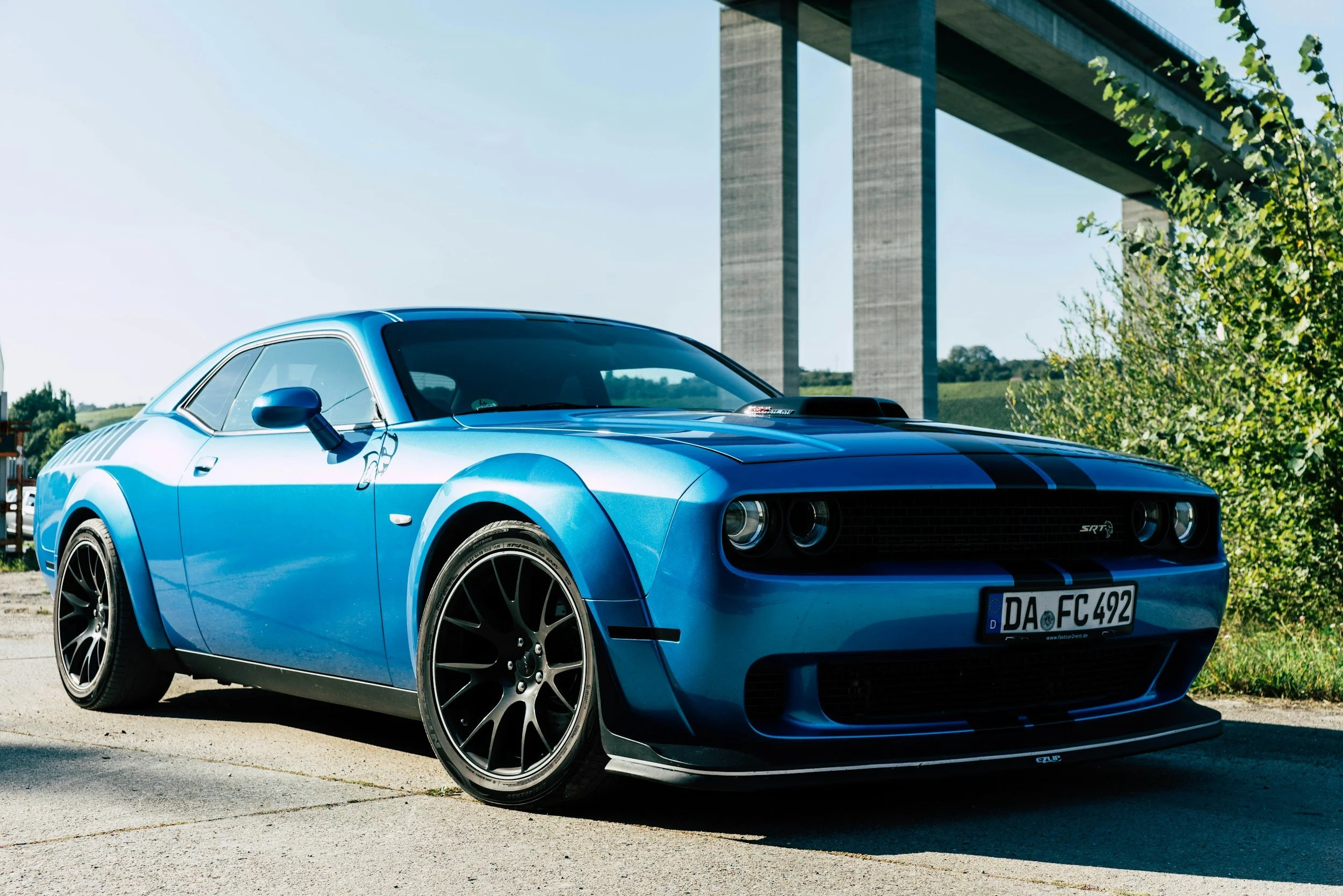 Blue Dodge Challenger SRT with black racing stripes parked outdoors with a bridge in the background.