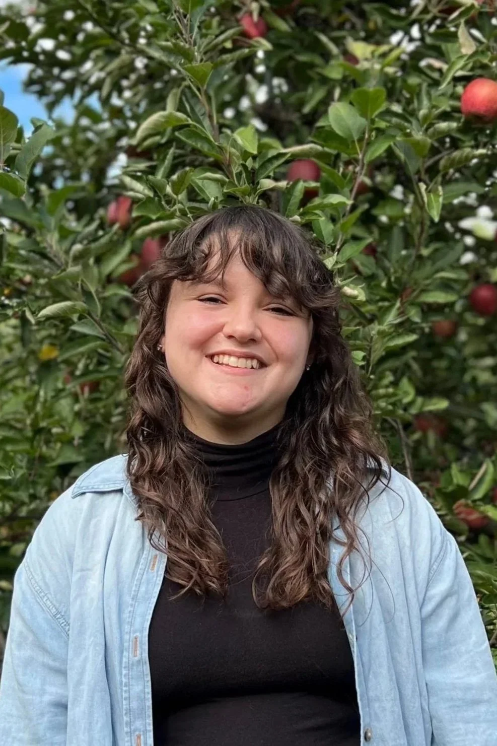 Photo of Emily, a white person with curly brown hair smiling and wearing an open denim button up with a black mock neck and apple trees behind them.