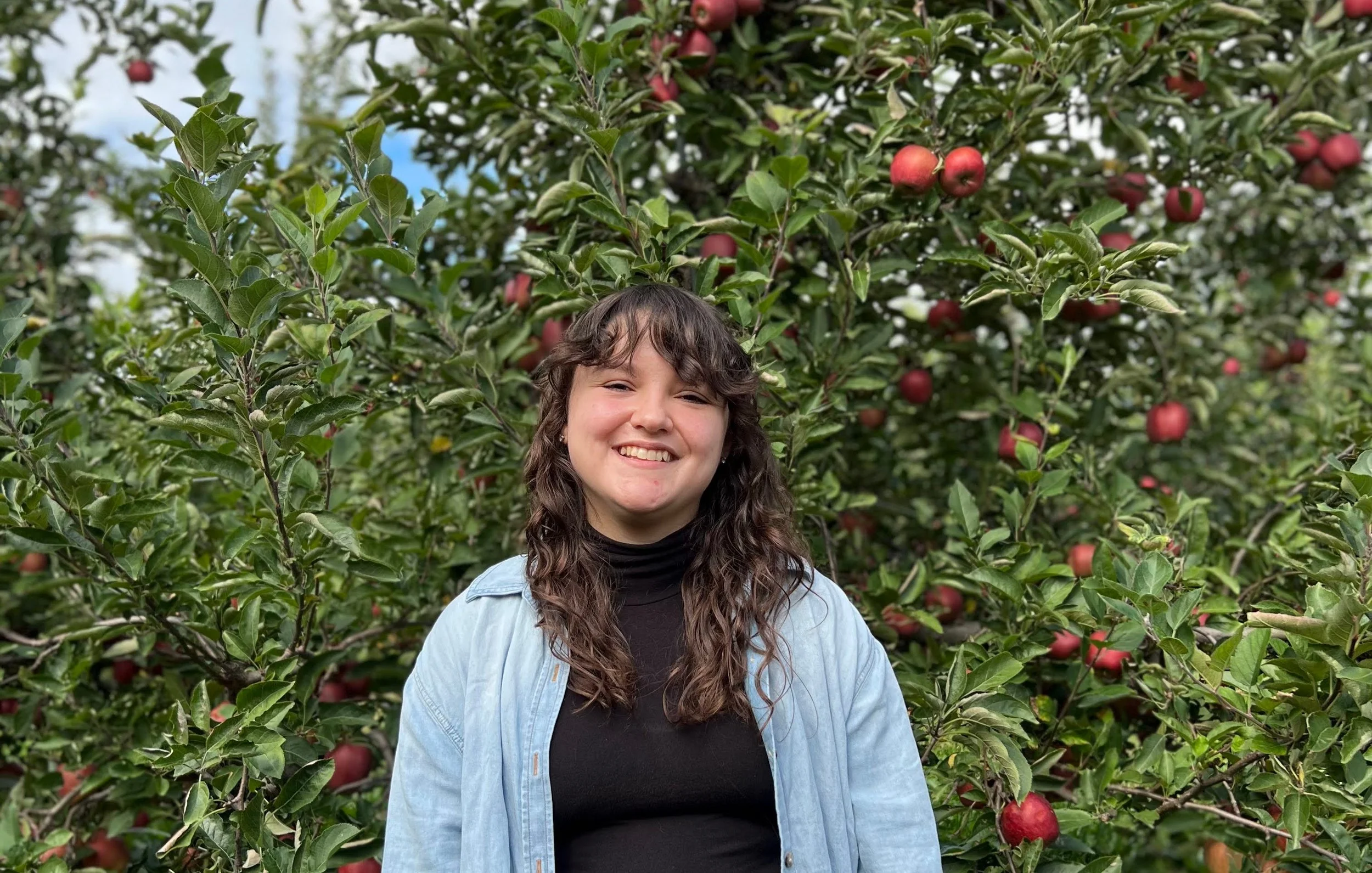 Photo of Emily, a white person with brown curly hair wearing a black shirt and open denim button up. They smile in front of some apple trees.