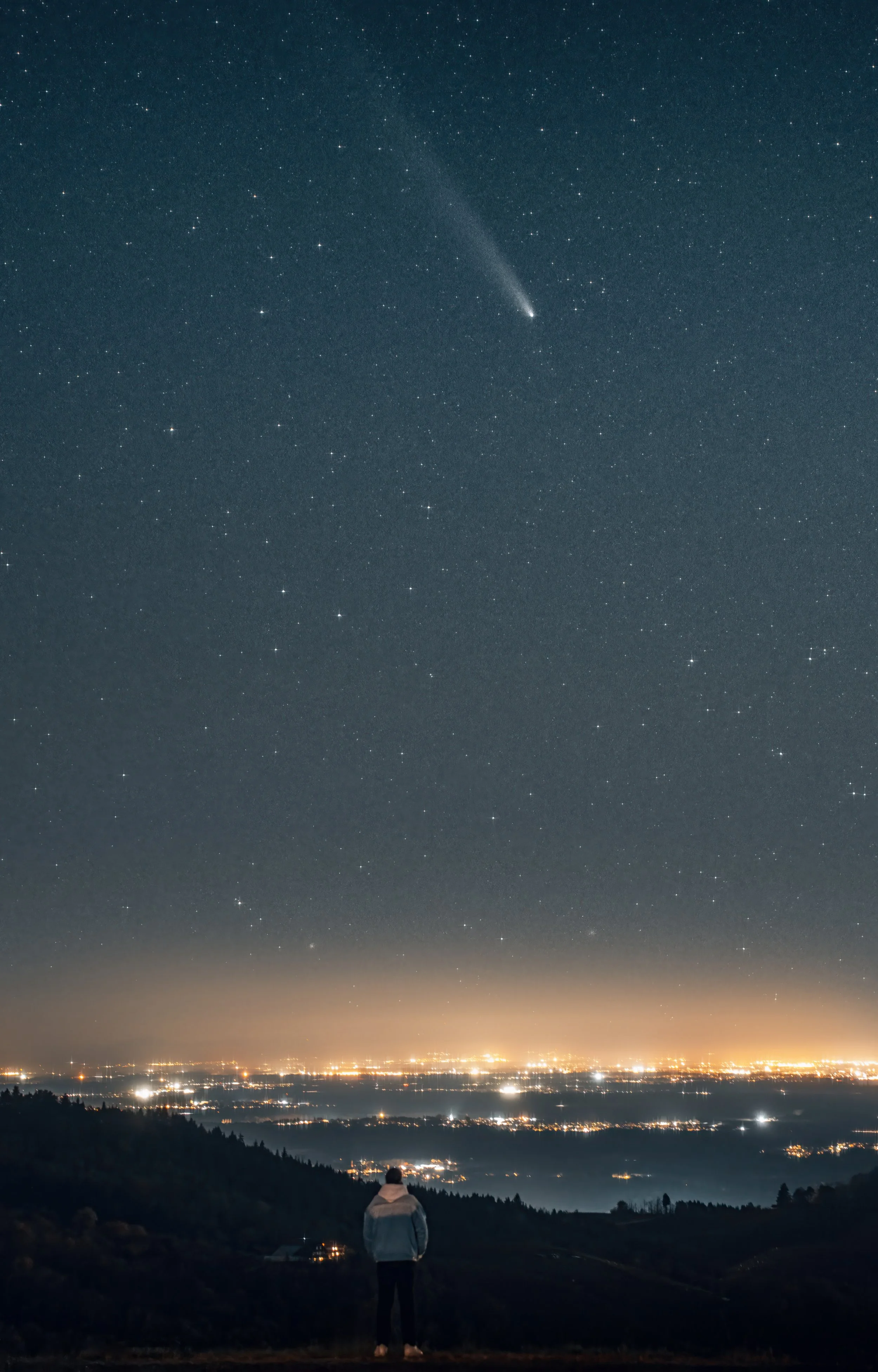 Ein Mensch steht auf einem Hügel und blickt auf eine Stadt mit Lichtern bei Nacht, Himmel voller Sterne und einen Kometen mit Schweif.