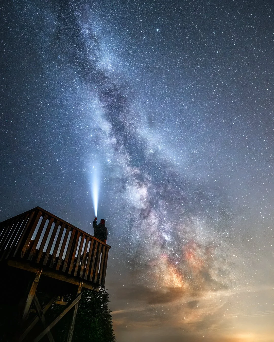 Eine Person steht auf einer Aussichtsplattform und leuchtet mit einer Taschenlampe in den Nachthimmel, voller Sterne und der Milchstraße.