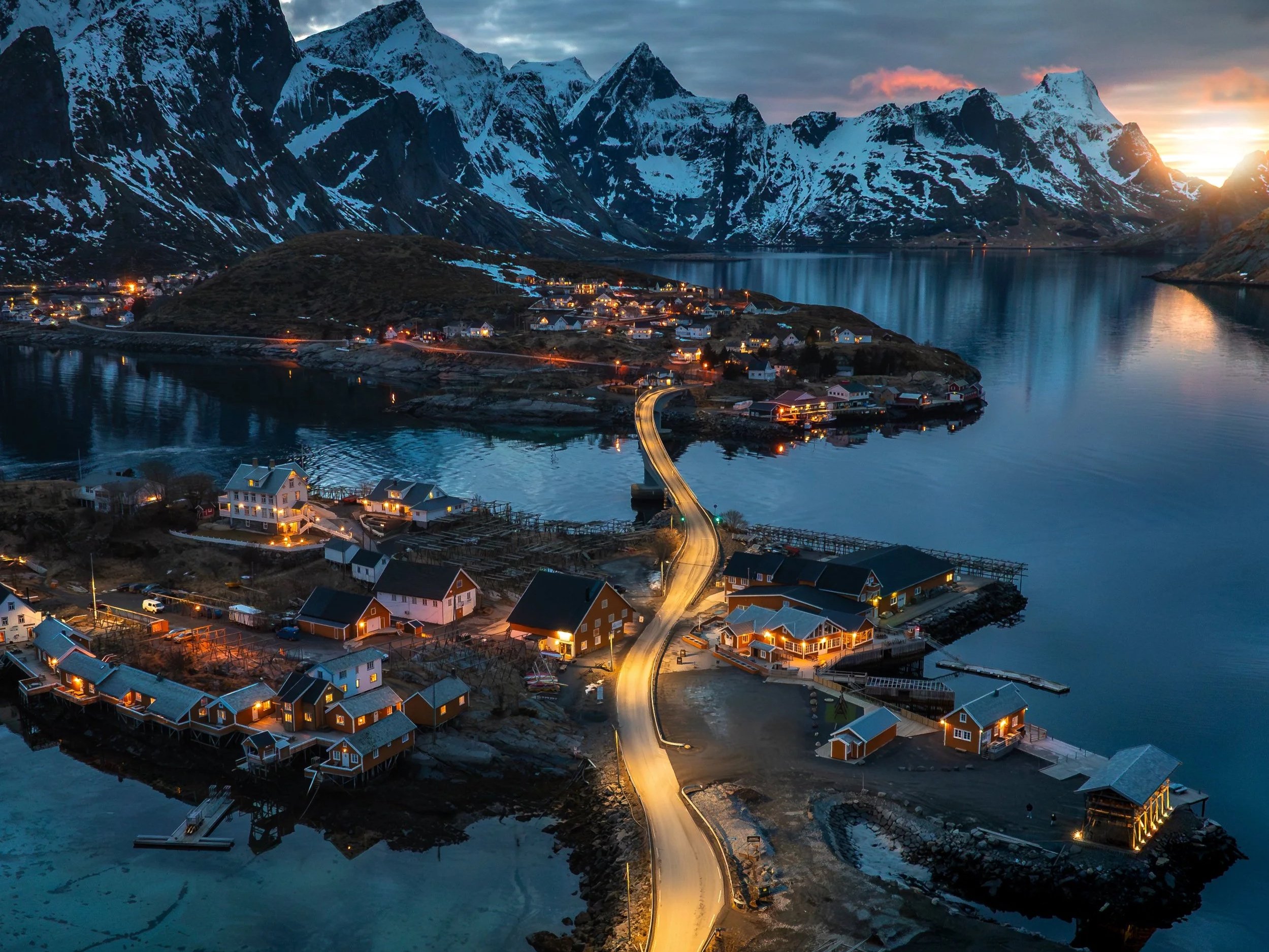 Landschaft bei Sonnenuntergang mit Bergen, Wasser und einem kleinen Dorf mit beleuchteten Häusern an der Küste.