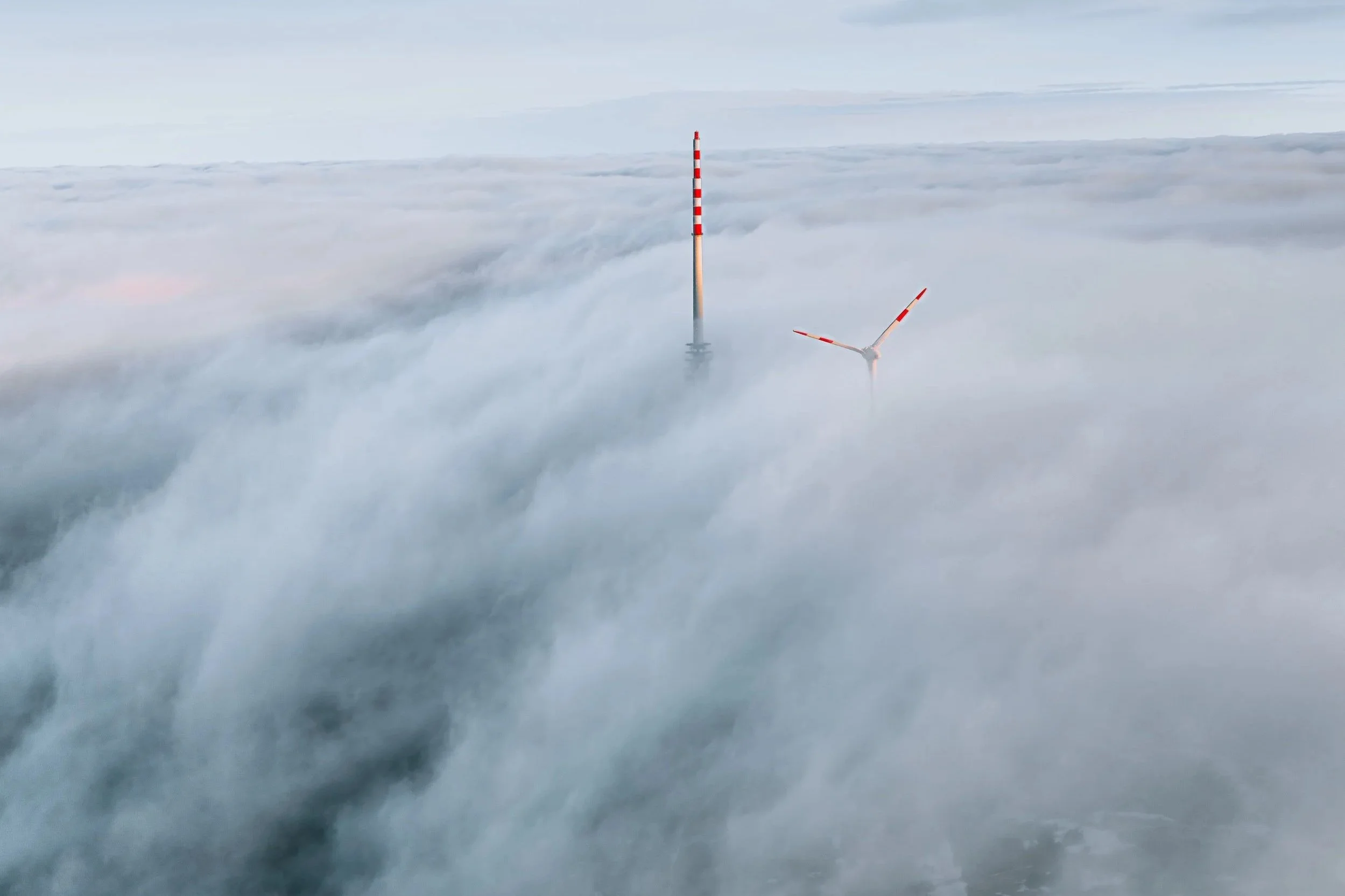 Windkraftanlage und Turm, die aus Wolken oder Nebel ragen, vor einem bewölkten Himmel.