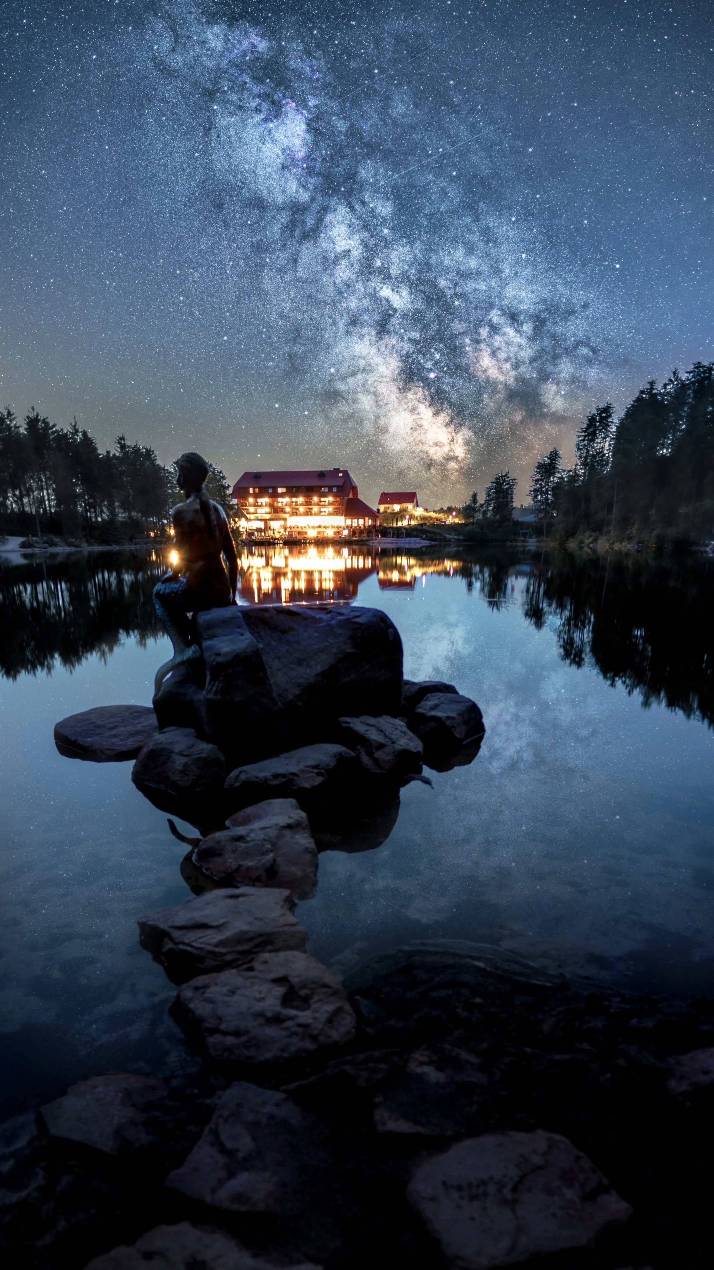 Sternenhimmel über einem ruhigen See mit einem sitzenden Jungen auf einem Stein und einem Gebäude am Ufer, das im Licht leuchtet.