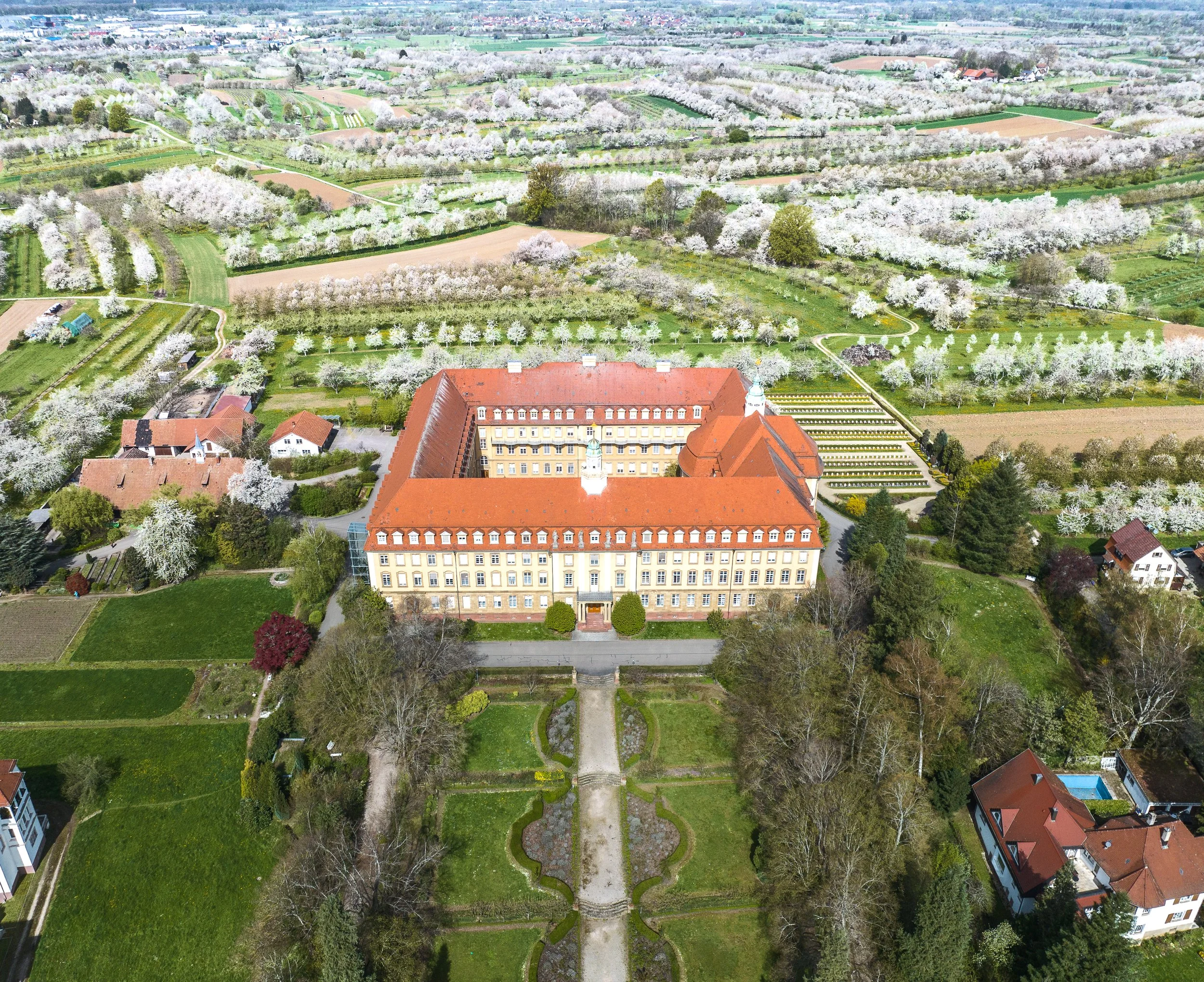 Luftaufnahme eines großen historischen Gebäudes mit rotem Dach, umgeben von Gärten, Bäumen und blühenden Obstbäumen in einer ländlichen Gegend mit Feldern im Hintergrund.