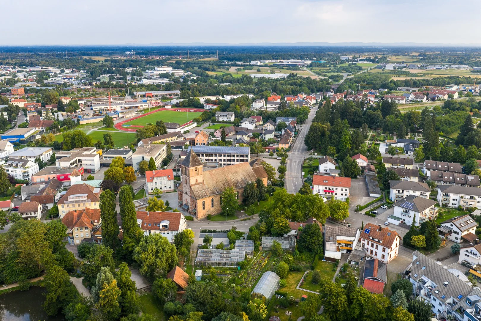 Luftaufnahme einer ruhigen Stadt mit Kirche, Wohnhäusern, Gärten, einem Sportplatz und umgebender Natur.