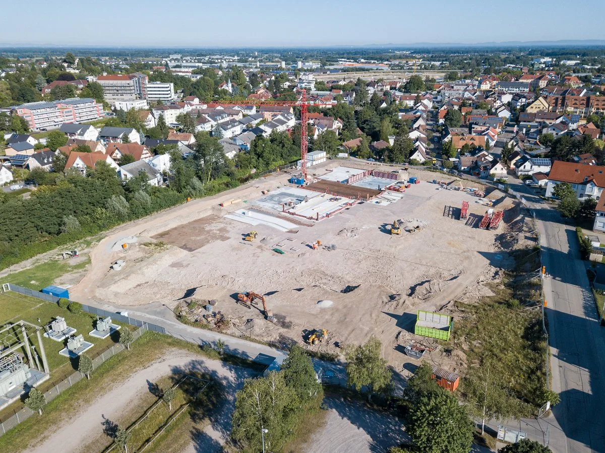Baustelle in einem Wohngebiet mit Baufahrzeugen und einem Baukran, umgeben von Häusern und Bäumen.