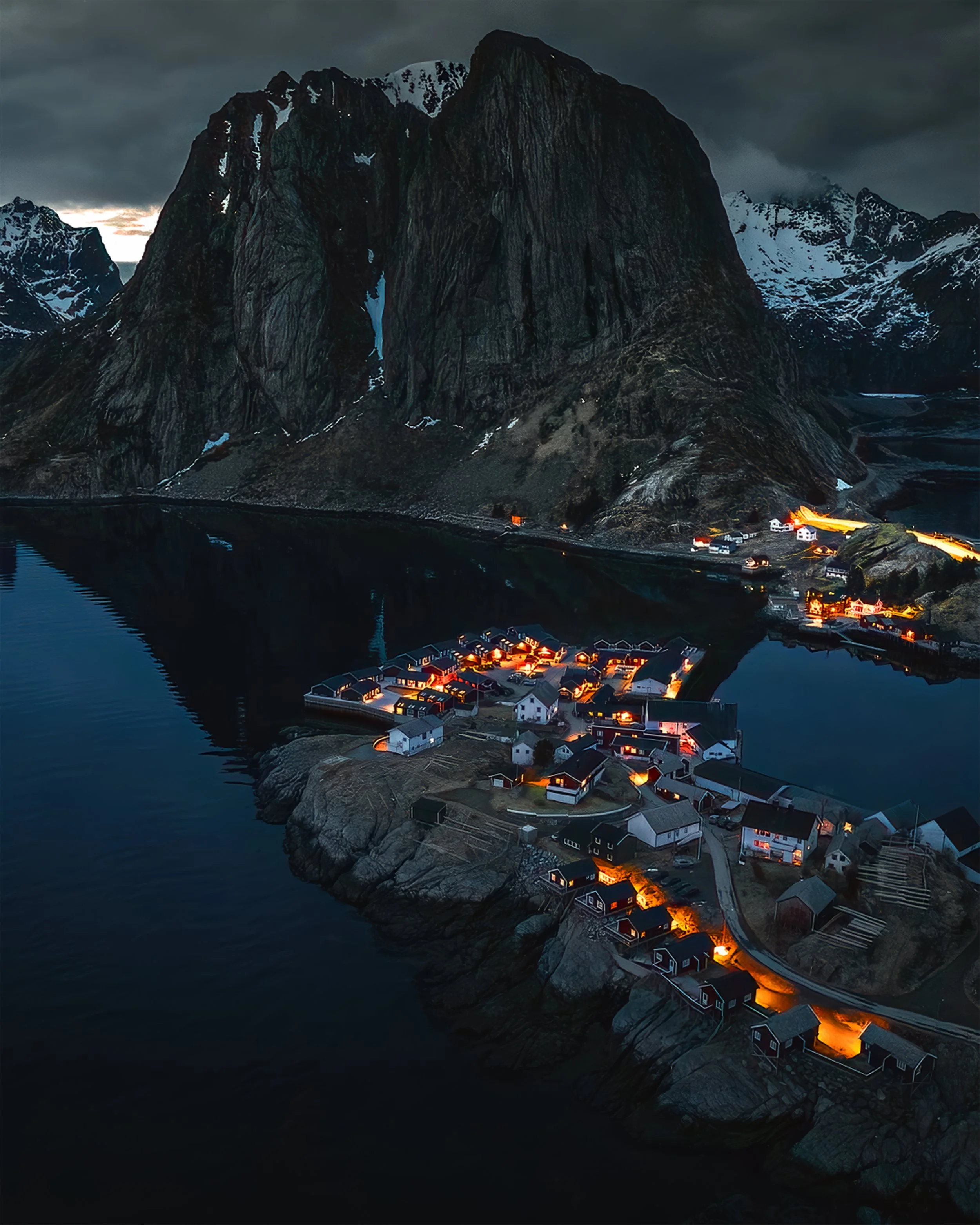 Abendliche Aufnahme eines Fjords mit einem kleinen Dorf, das an den Ufern und auf den Felsen liegt, vor einer beeindruckenden Bergsilhouette im düsteren Himmel.