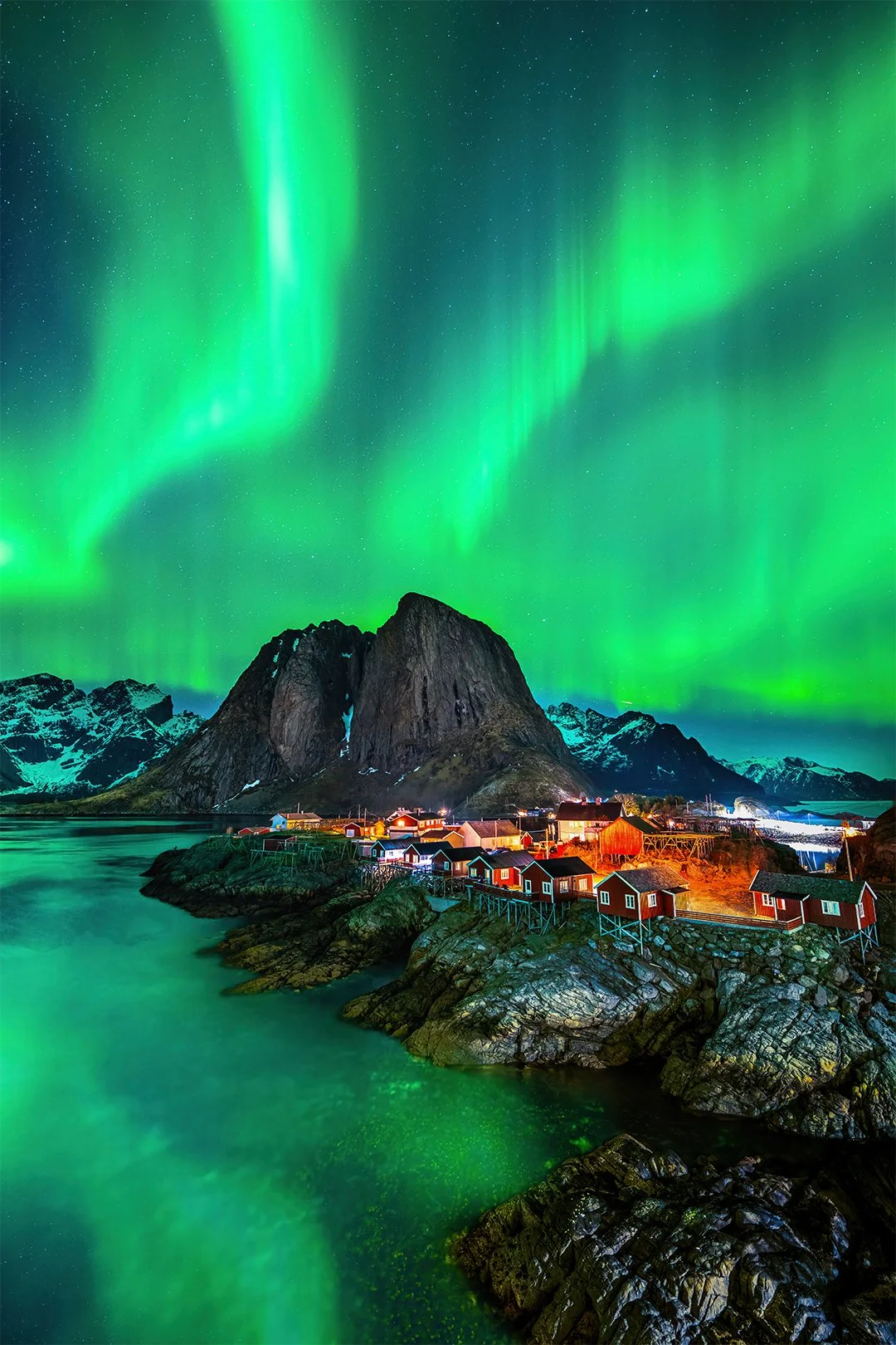 Nordlichter leuchten grün am Himmel über einer dänischen Fischerkoje an der norwegischen Küste mit Bergen im Hintergrund.